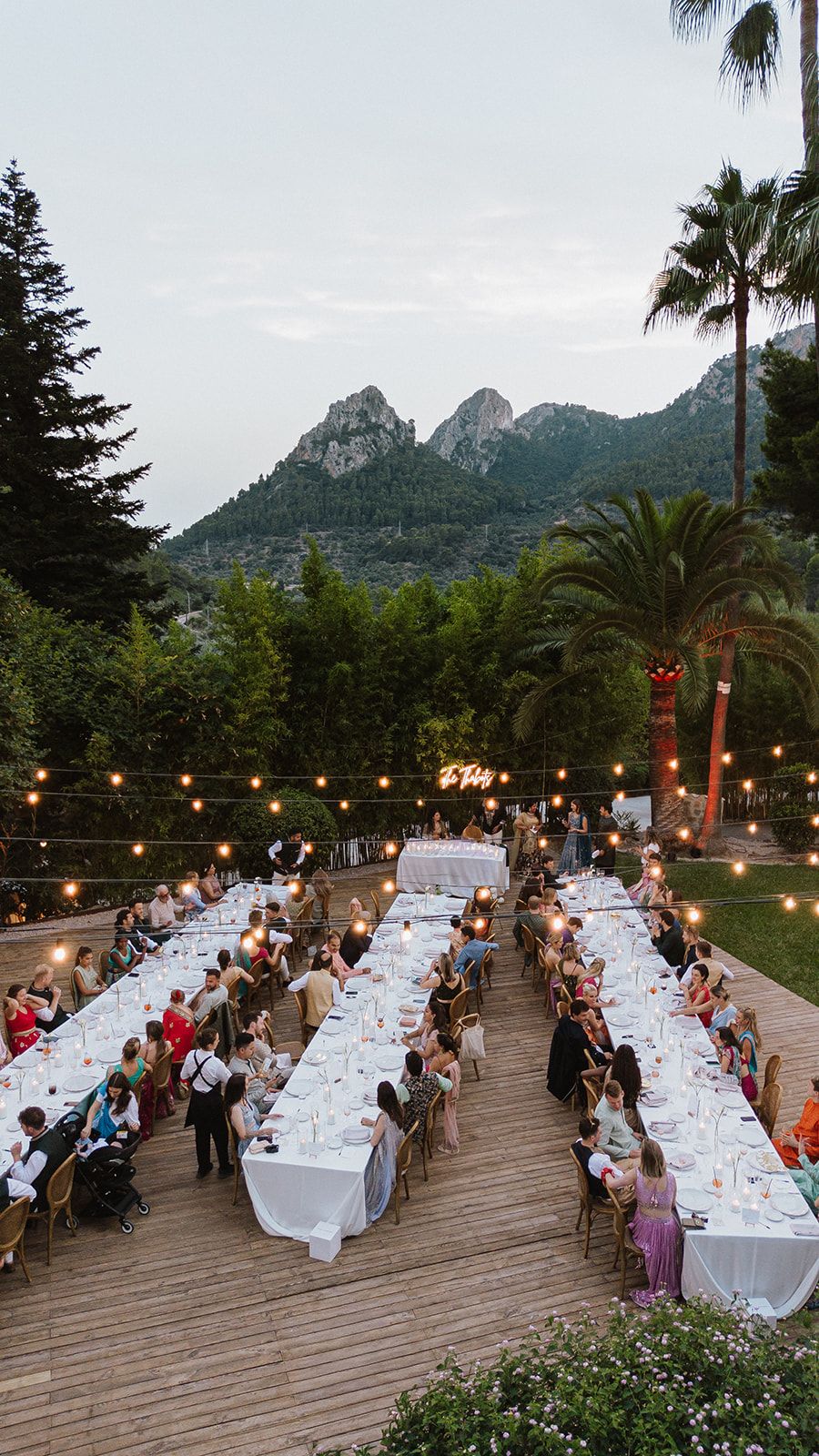 Aerial view of elegant outdoor wedding reception with long tables, string lights, and mountain backdrop.