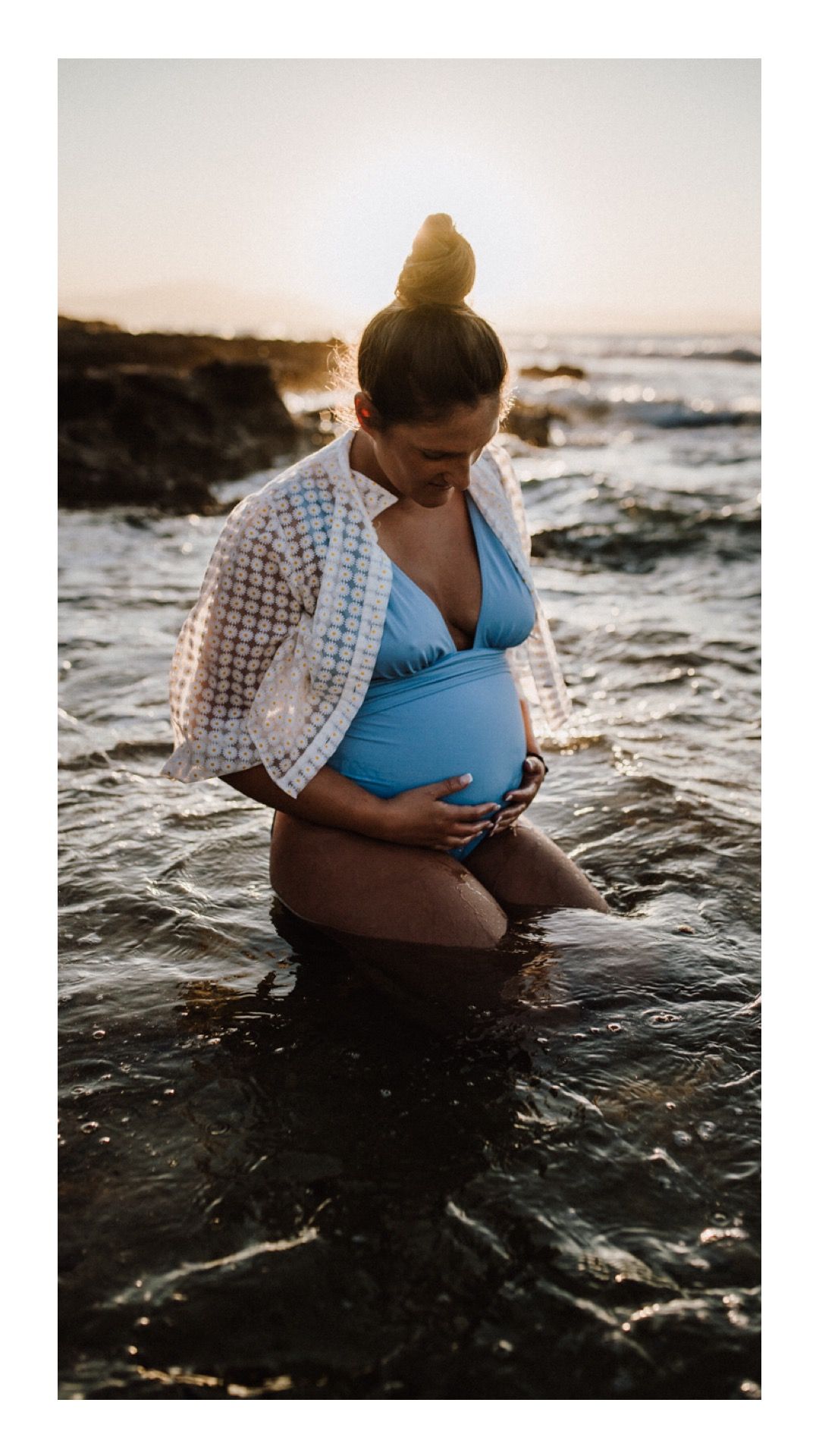 Pregnant woman in blue bikini and white cover-up cradles her belly while standing in ocean water at sunset.