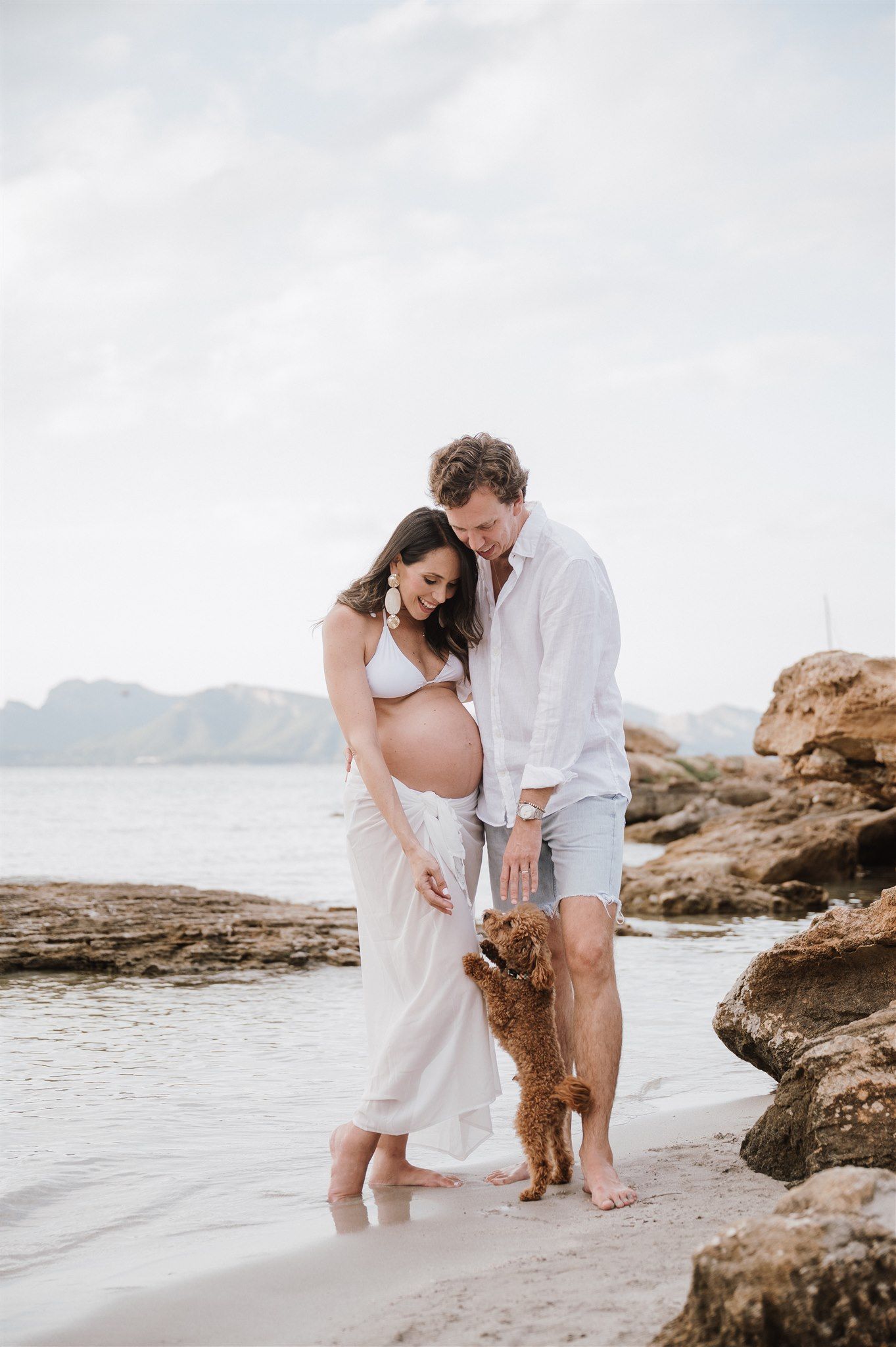 Pregnant couple in white outfits smiling at their golden doodle puppy on a rocky beach with mountains in the background.