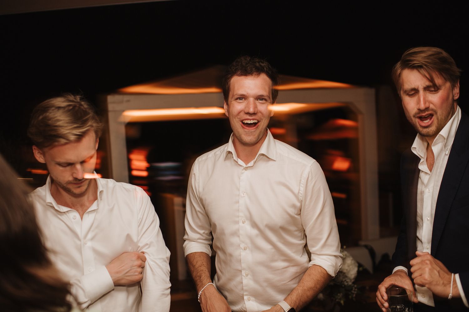 Three men in dress shirts laugh and celebrate together at a wedding evening reception.