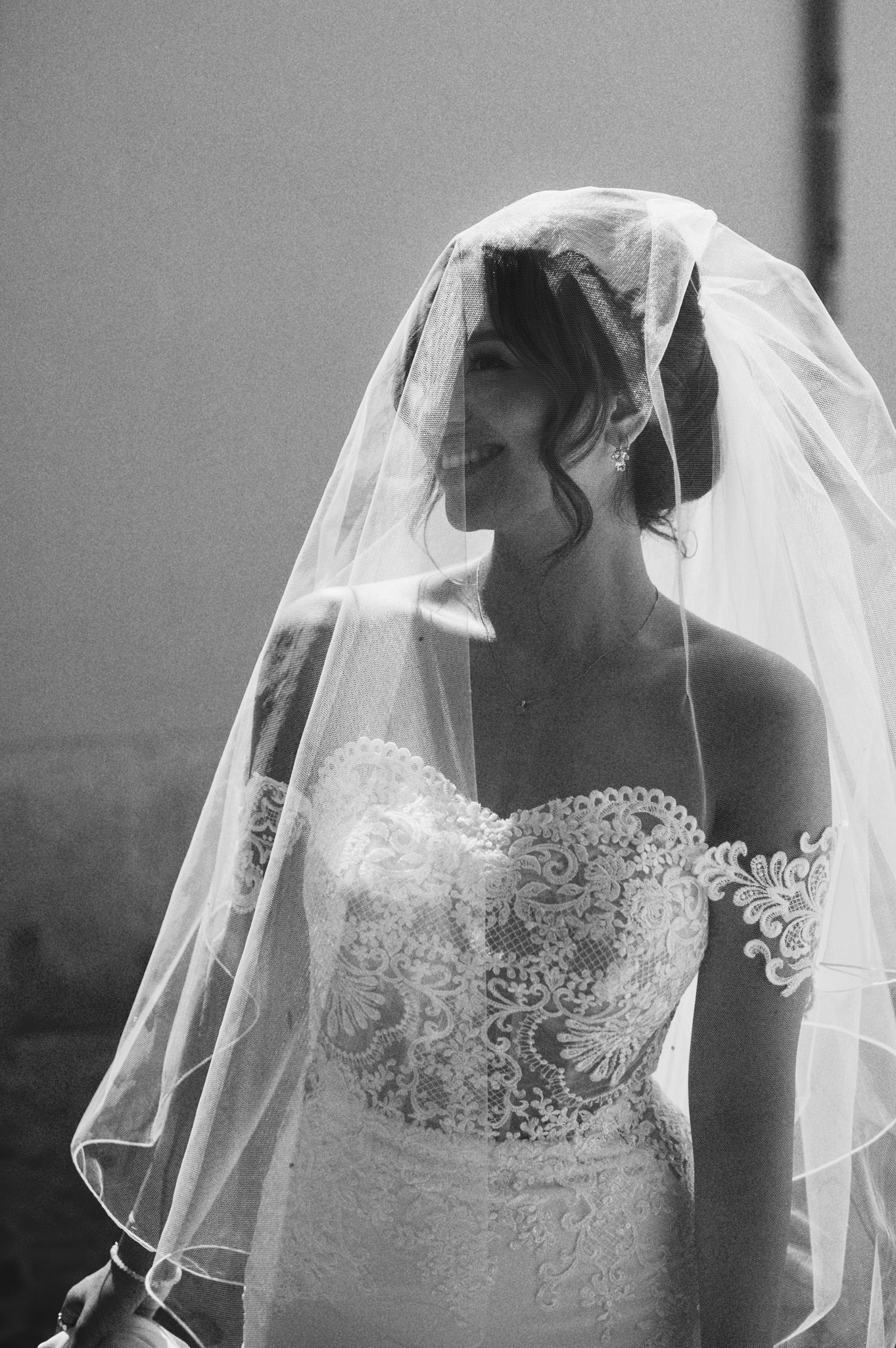 Black and white portrait of smiling bride wearing lace strapless wedding dress with flowing veil draped over head.
