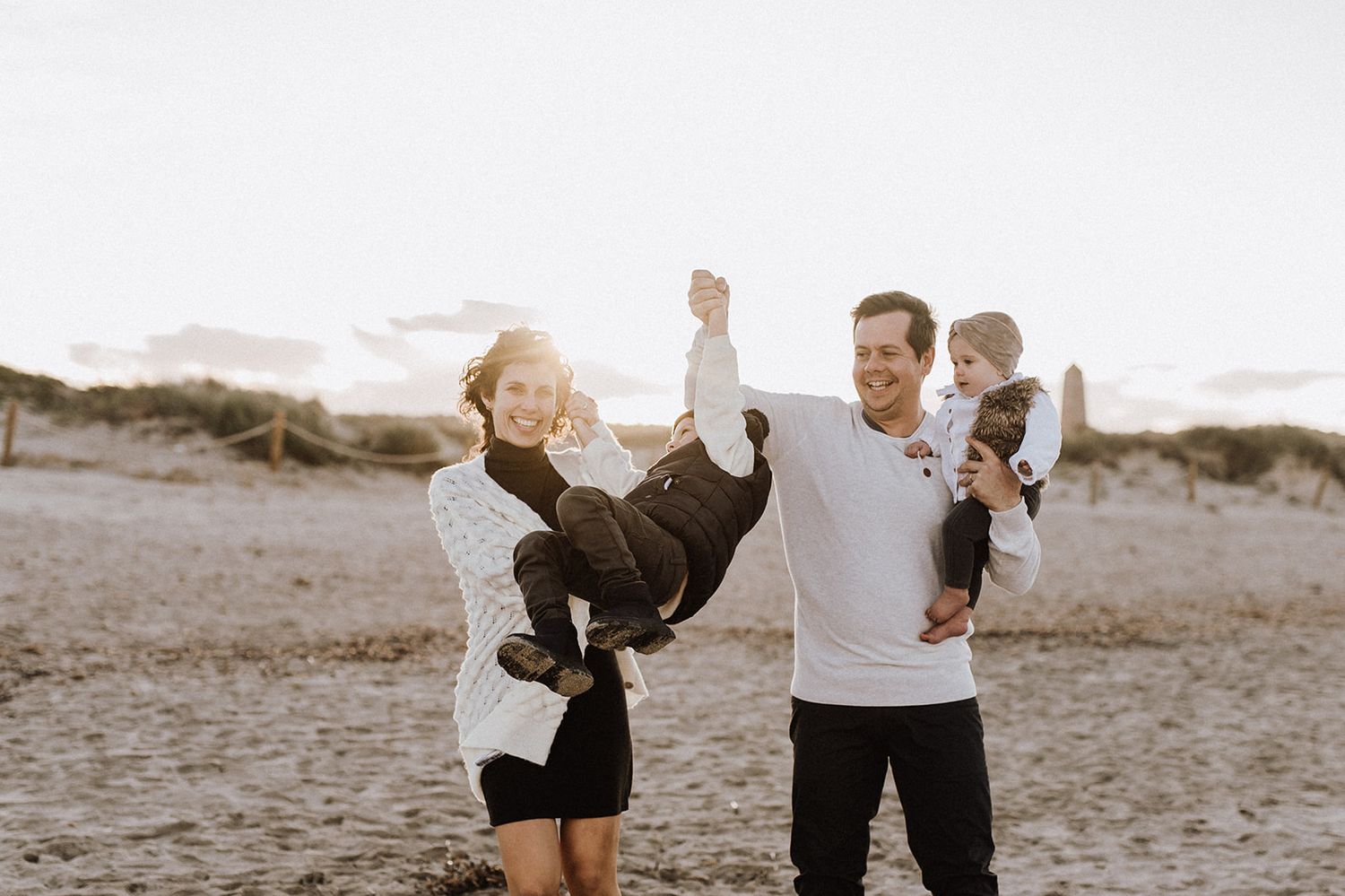 Young family laughing and playing with two babies on a sandy beach at golden hour.