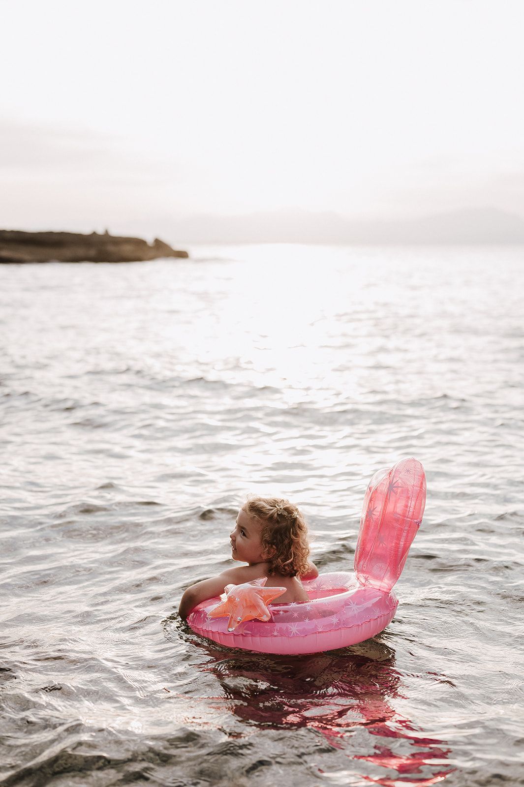 Young girl floating in pink flamingo inflatable ring in calm ocean water at dusk.