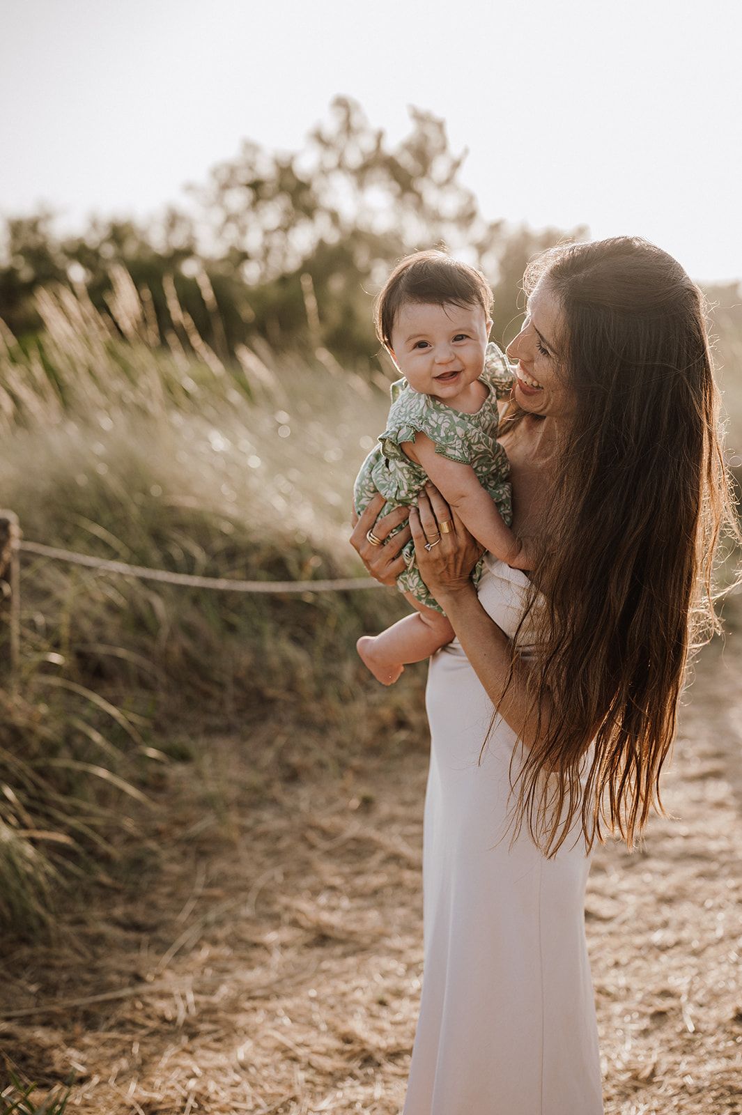 A mother holds her smiling baby outdoors in golden sunlight near tall grasses.