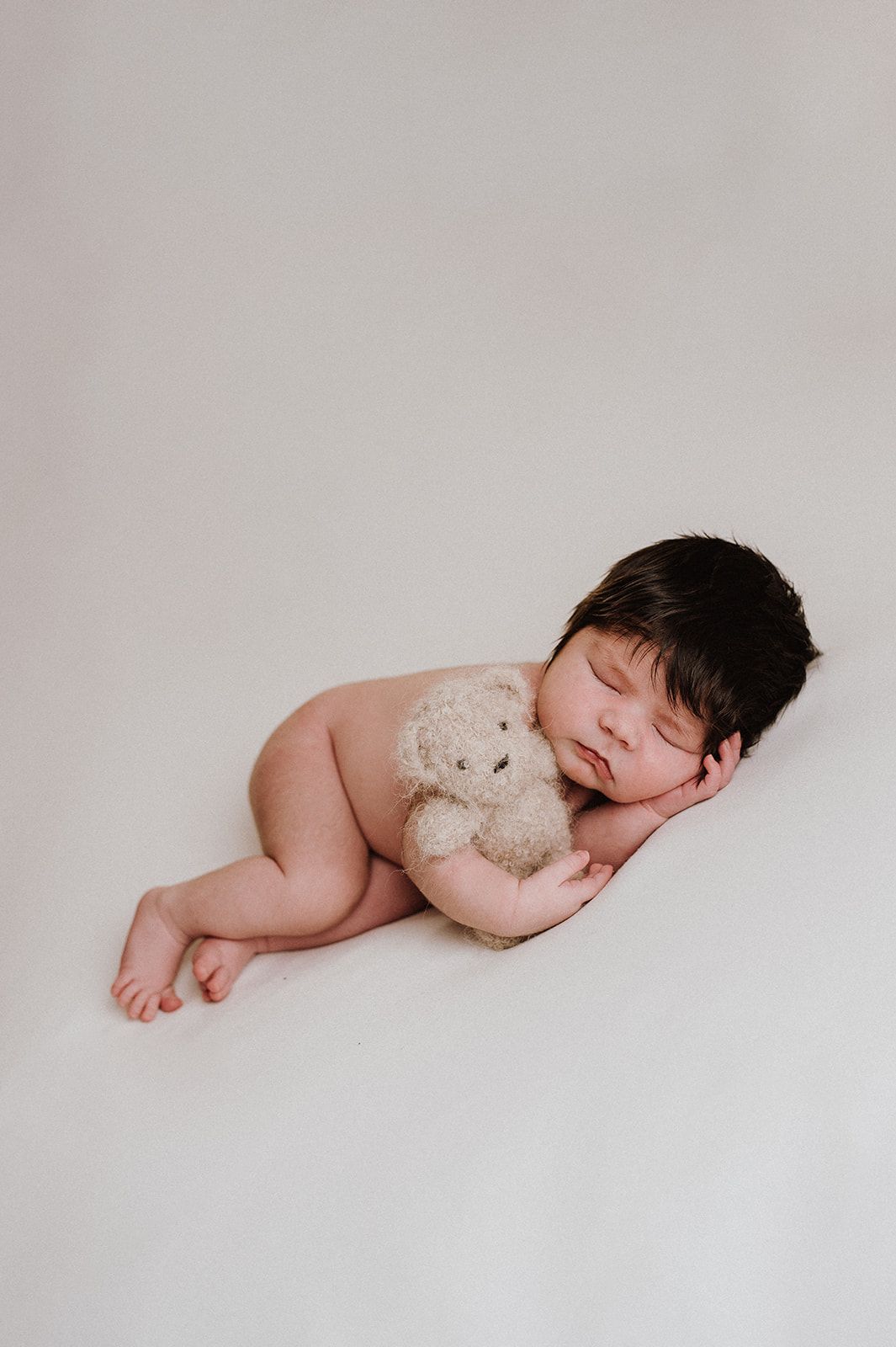 Sleeping newborn baby with dark hair cuddling a small teddy bear on a white background.