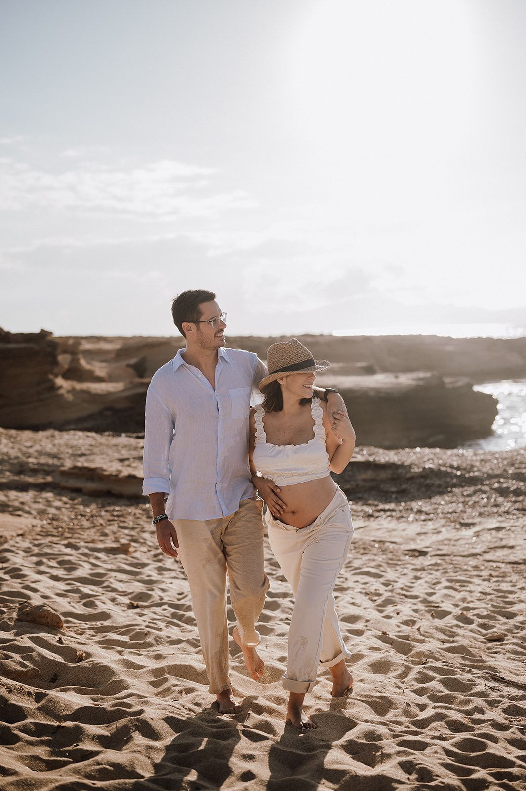 Couple walking on sandy beach, pregnant woman in white outfit and hat, man in light blue shirt, sunny coastal backdrop.
