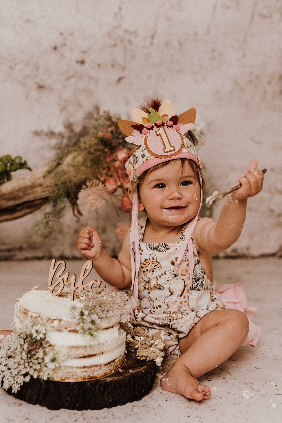 Baby girl celebrating her first birthday with a cake smash, wearing a floral crown with number 1.