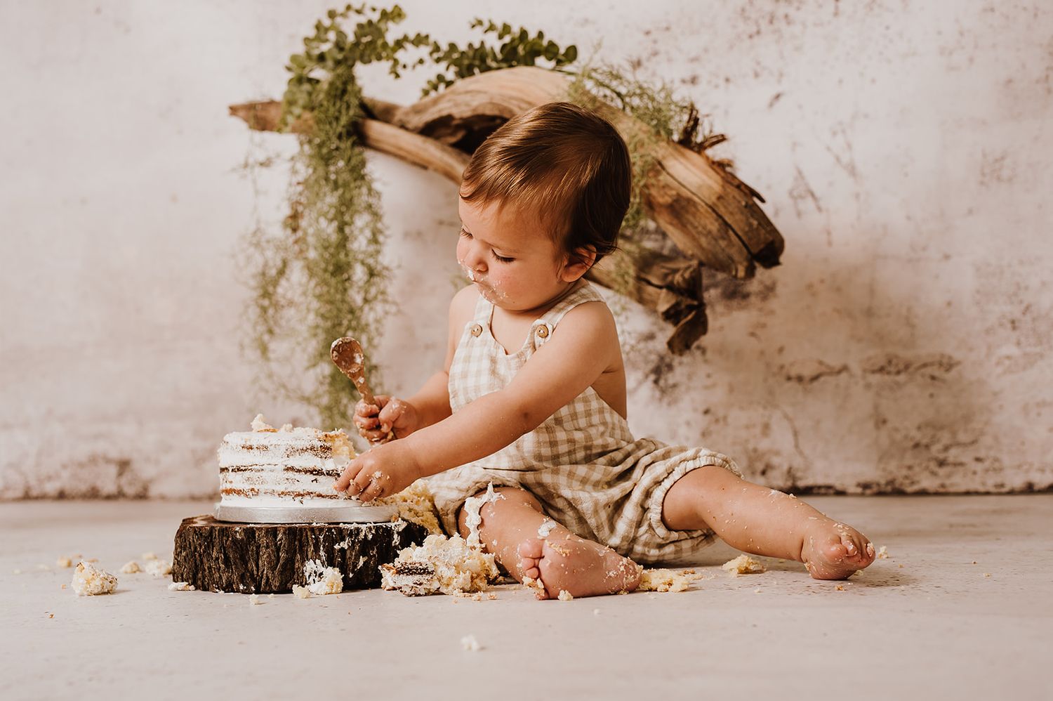 Toddler in gingham romper sitting on floor touching a small cake on a wood stump with greenery backdrop.