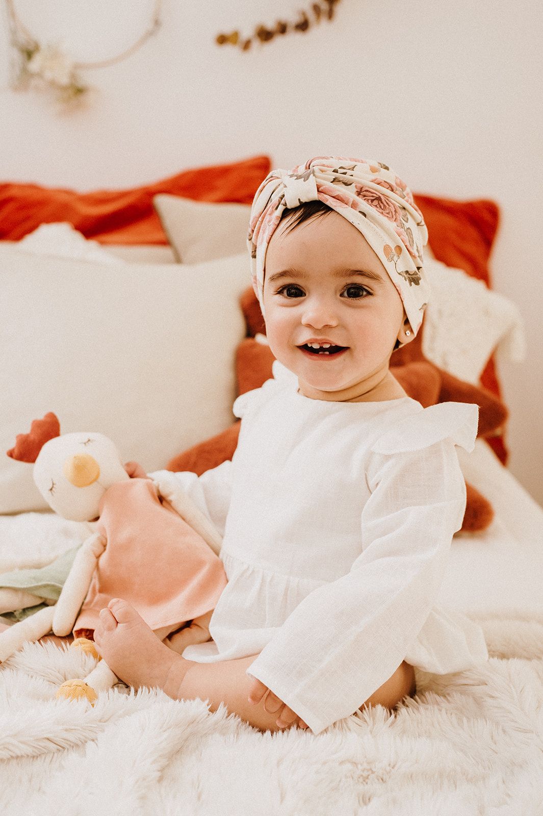 Smiling baby girl in white dress and floral turban headband sitting on bed holding a soft toy doll.