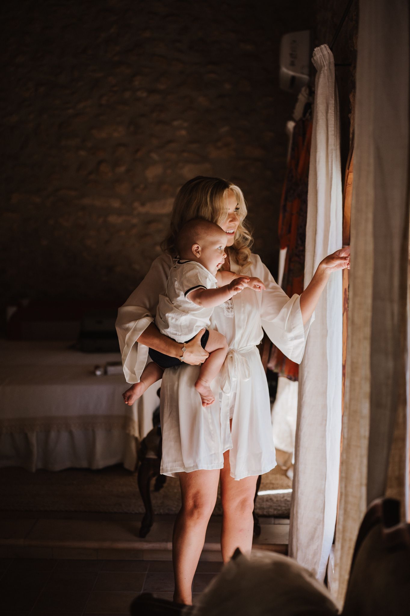 Blonde woman in white silk robe holding a baby near a sunlit curtain in a rustic stone-walled room.