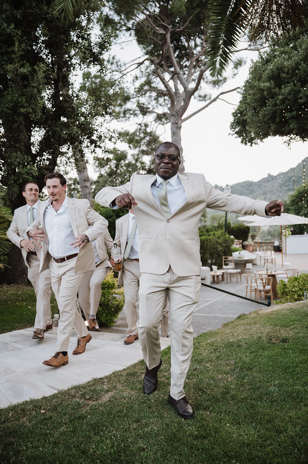 Groomsmen in beige suits walking energetically through garden at outdoor wedding reception with palm trees in background.
