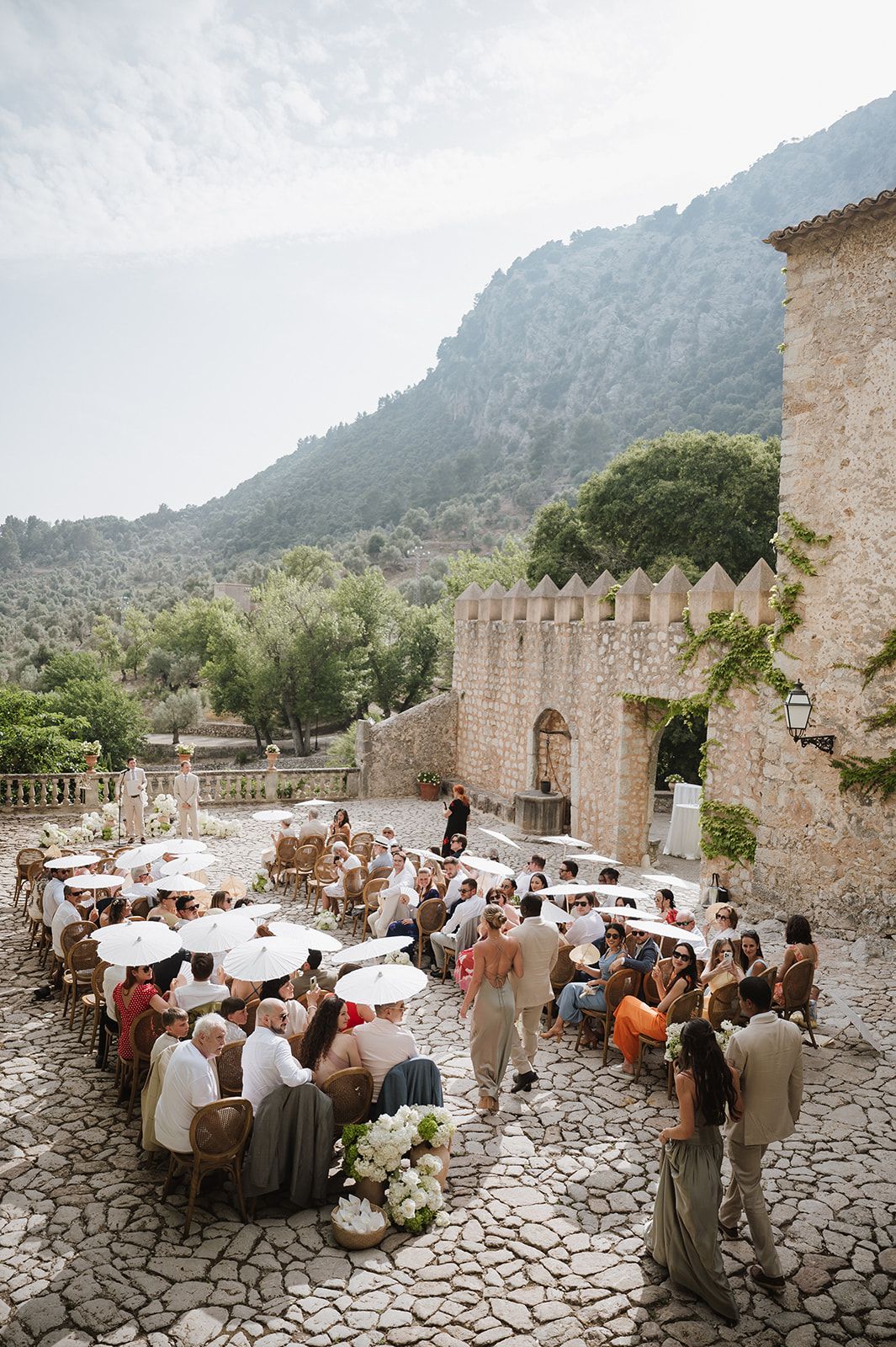 Aerial view of outdoor wedding ceremony at stone castle courtyard with guests seated under white parasols in mountains.