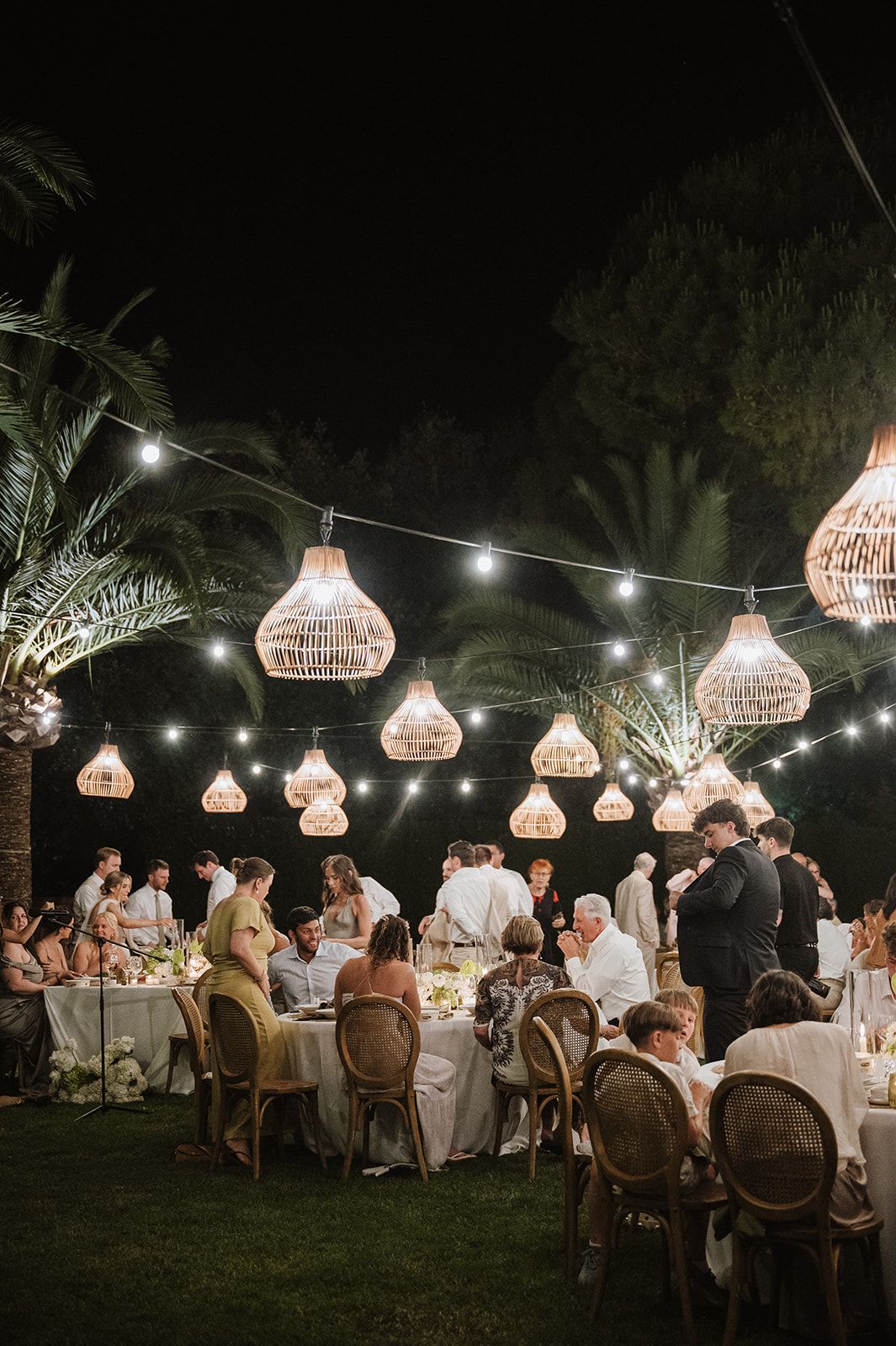 Elegant outdoor wedding reception at night with glowing rattan pendant lights and string lights over dining guests.