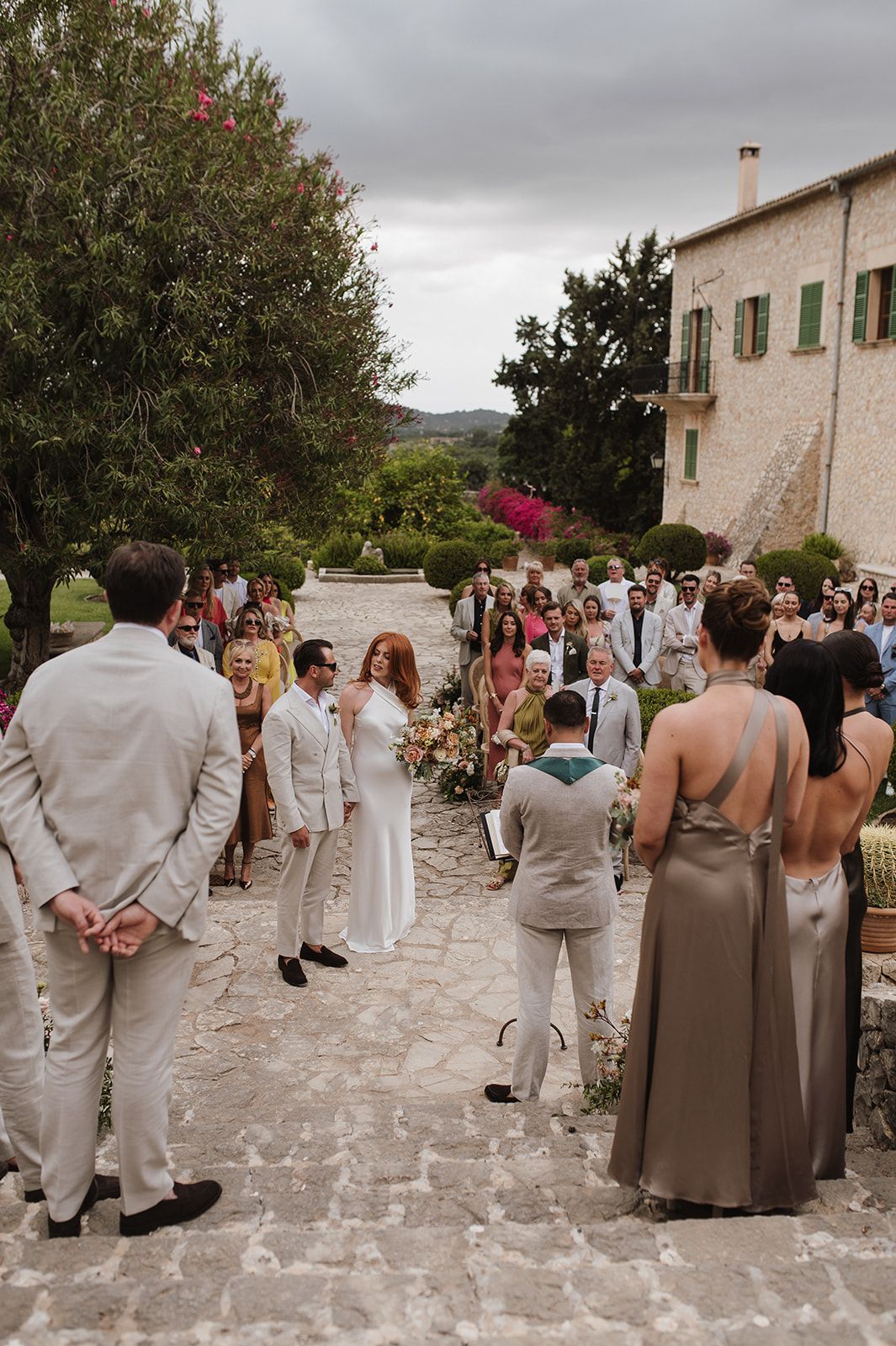 Wide shot of outdoor wedding ceremony at a rustic stone villa with guests lining the aisle.