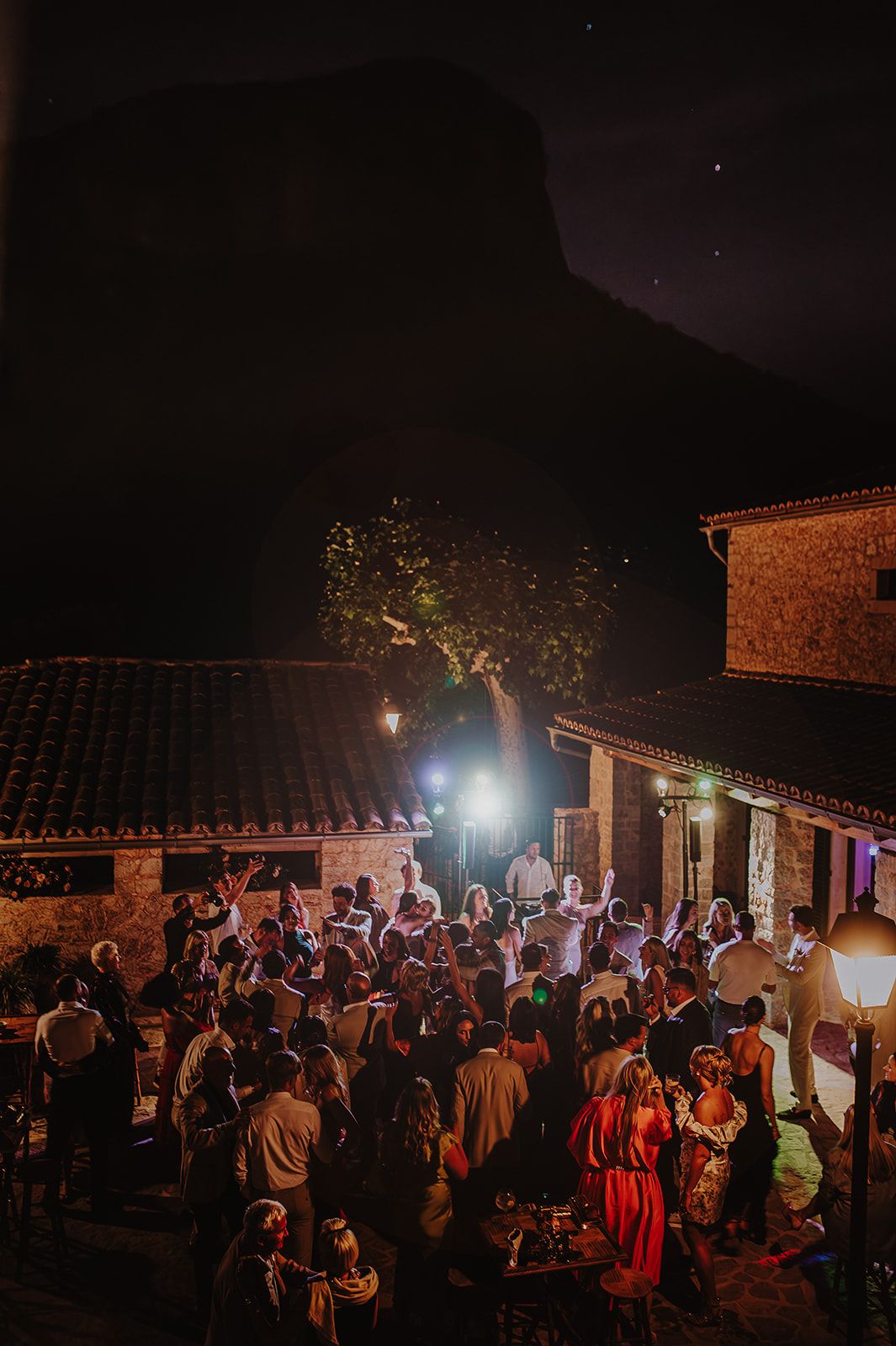 Aerial view of wedding guests dancing outdoors at night with a dramatic rocky mountain backdrop.