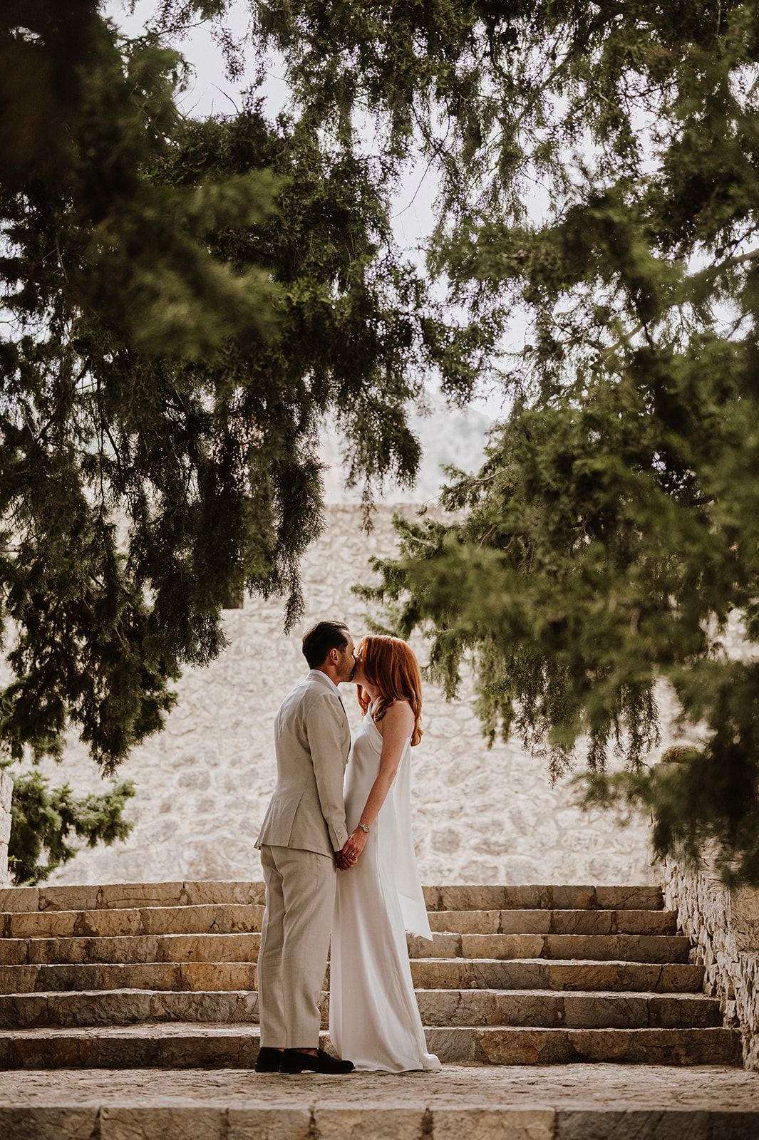 Bride and groom sharing a romantic kiss on stone steps framed by lush overhanging cypress trees.
