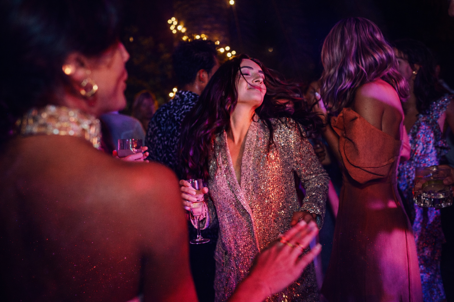 Women in elegant dresses dancing and socializing at a glamorous evening party with pink lighting.