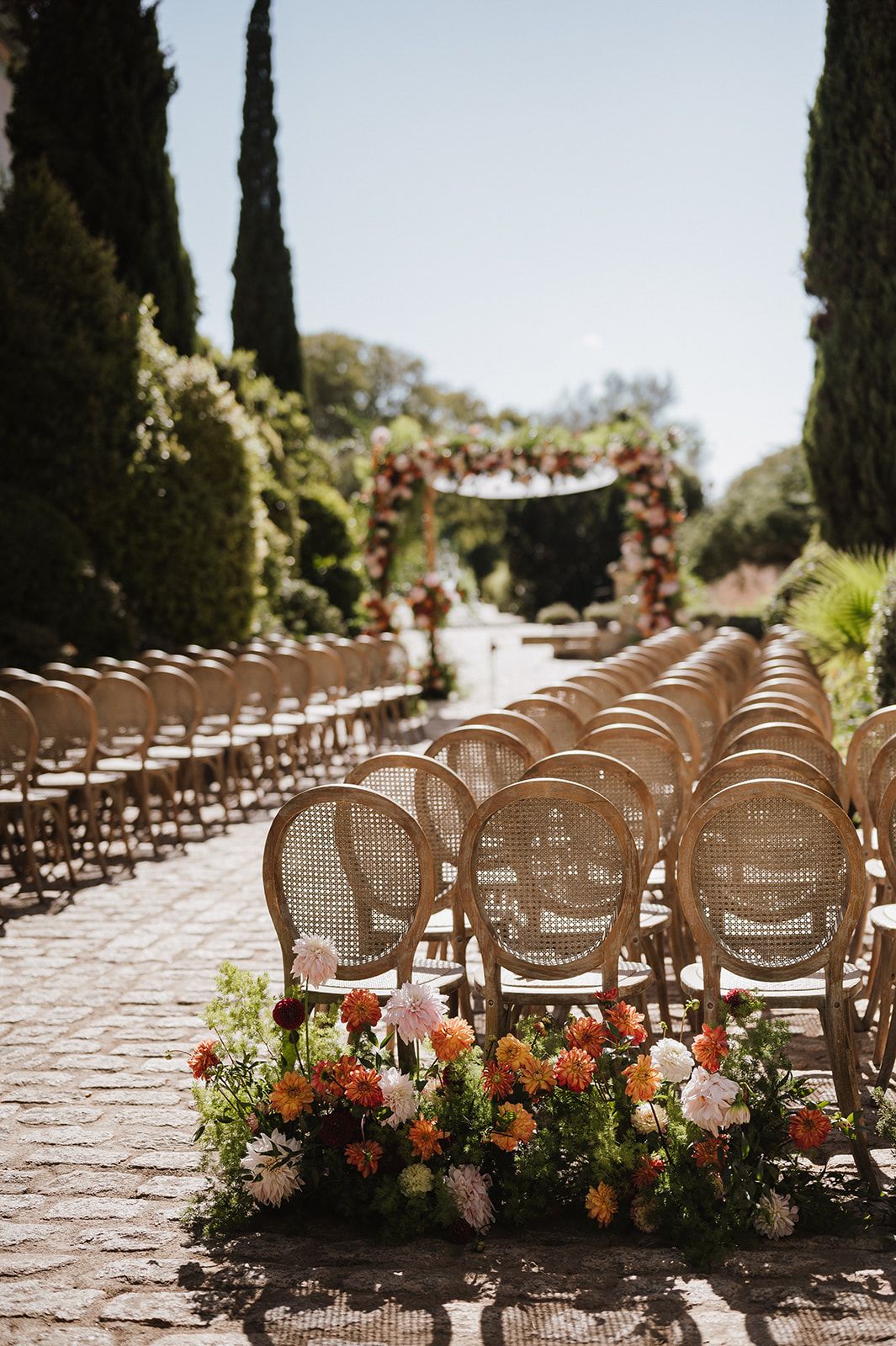 Rows of rattan chairs line a cobblestone aisle with colorful floral arrangements leading to a floral arch at an outdoor wedding.