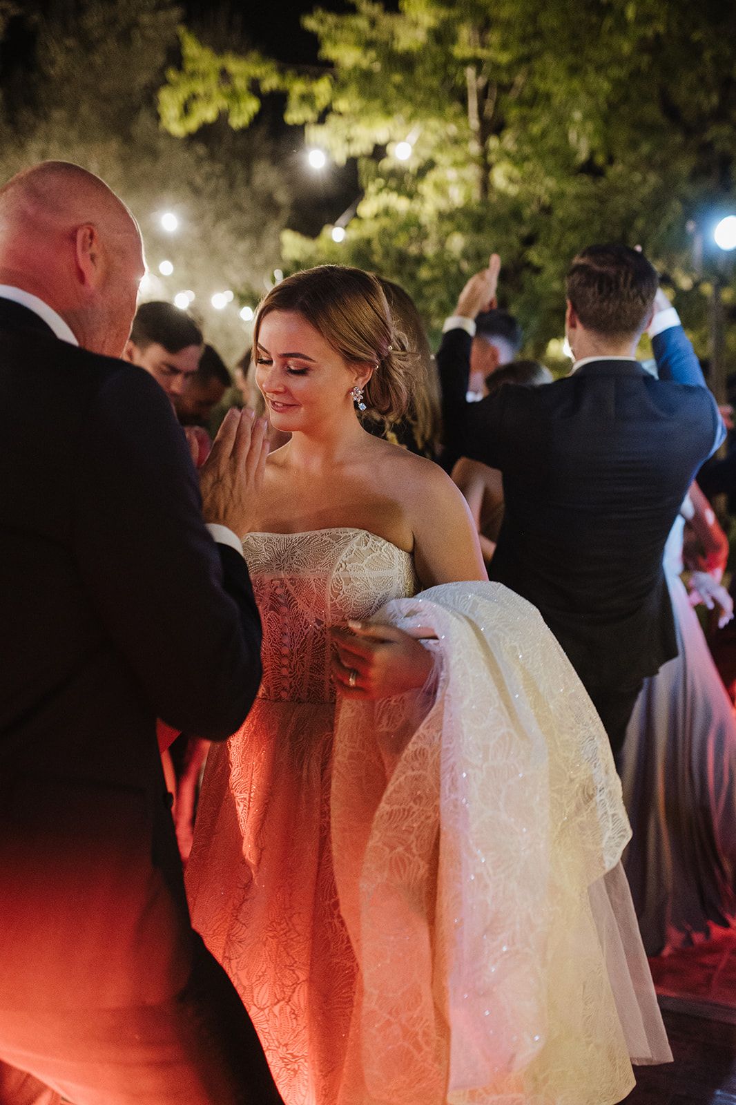 Bride in strapless embellished gown dancing joyfully at outdoor evening wedding reception.