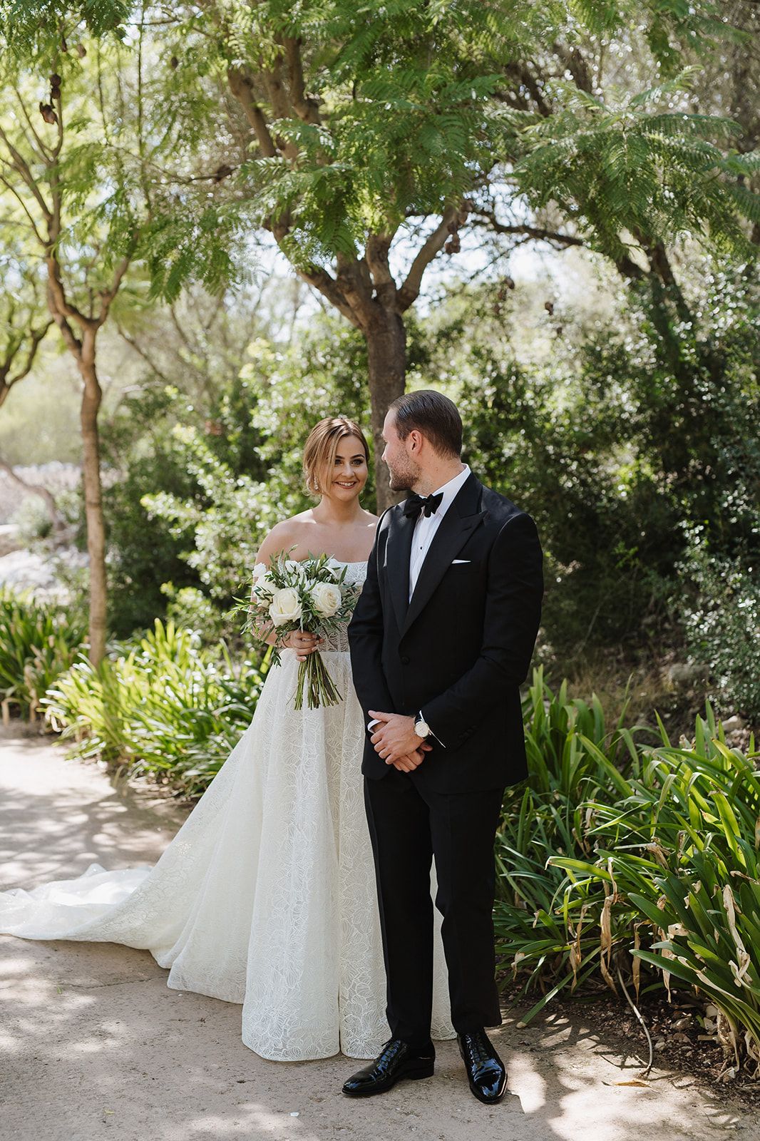 Bride in lace gown and groom in black tuxedo share a romantic moment in a lush garden setting.