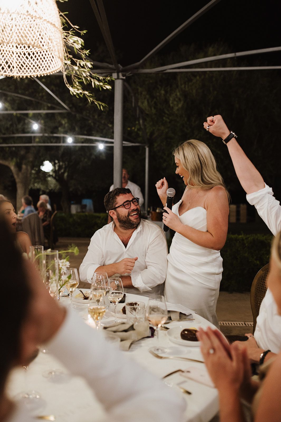 Joyful bride gives speech at outdoor wedding reception while groom laughs at decorated dinner table.