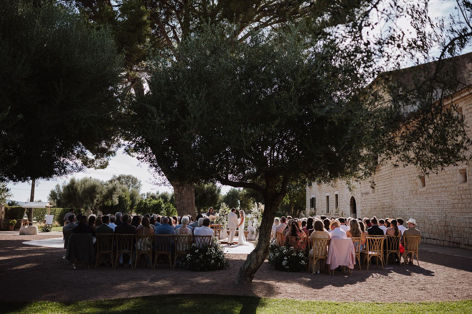 Guests seated at outdoor wedding ceremony beneath large shade trees.