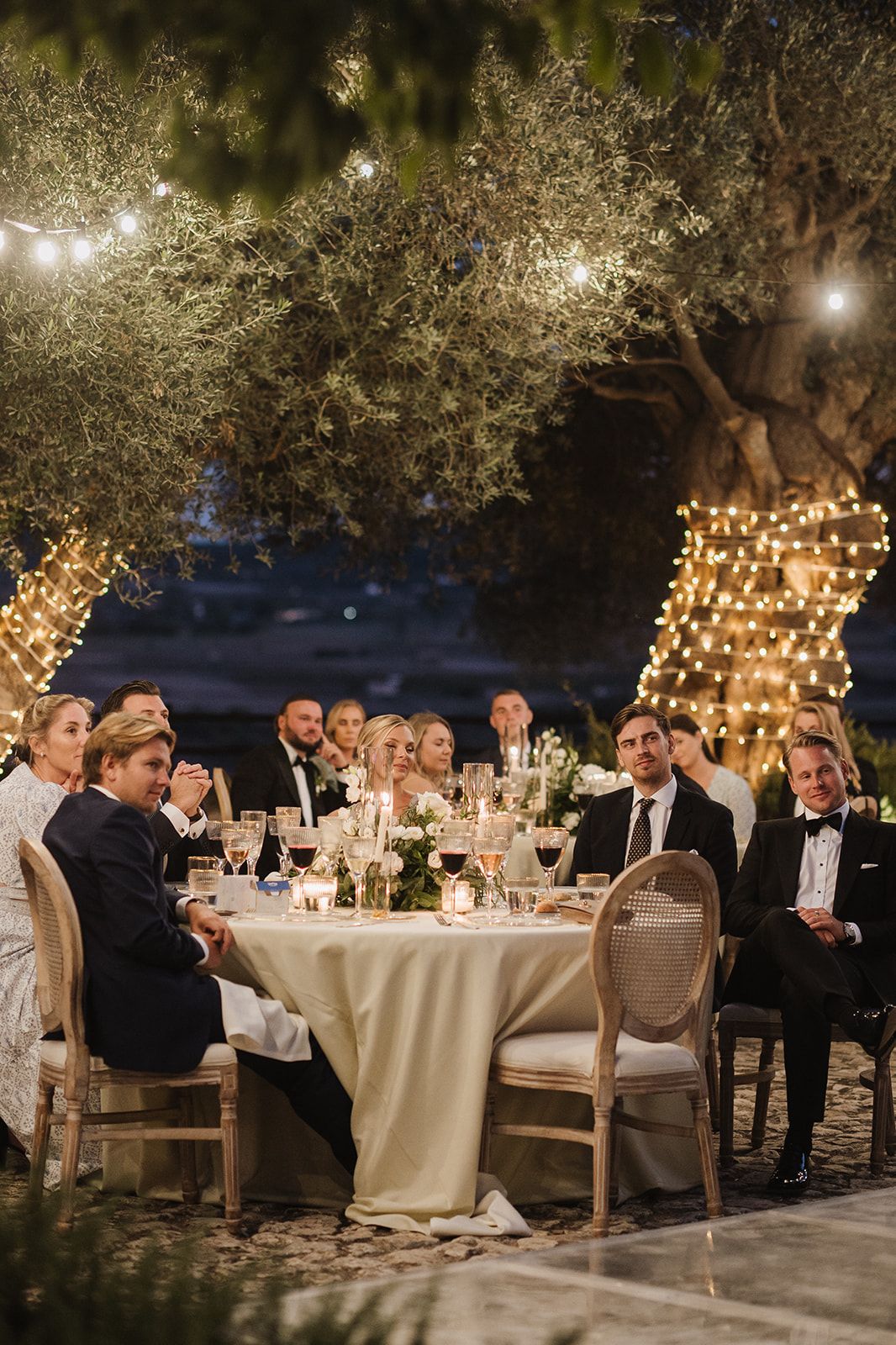 Wedding guests in formal attire dine at candlelit tables beneath fairy-light-wrapped olive trees at night.