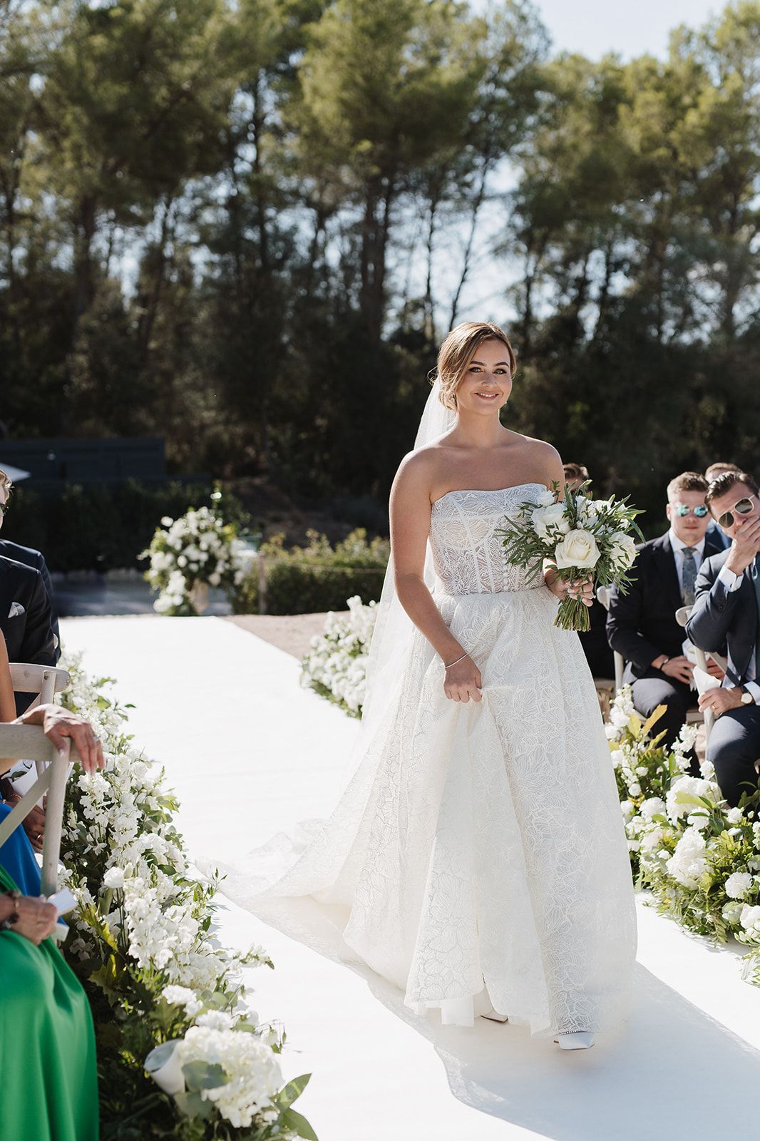 Radiant bride walks down white aisle in strapless lace gown holding a white floral bouquet outdoors.