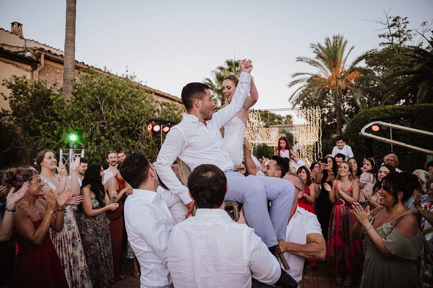 Bride and groom lifted by guests during lively hora dance at outdoor wedding reception at sunset.