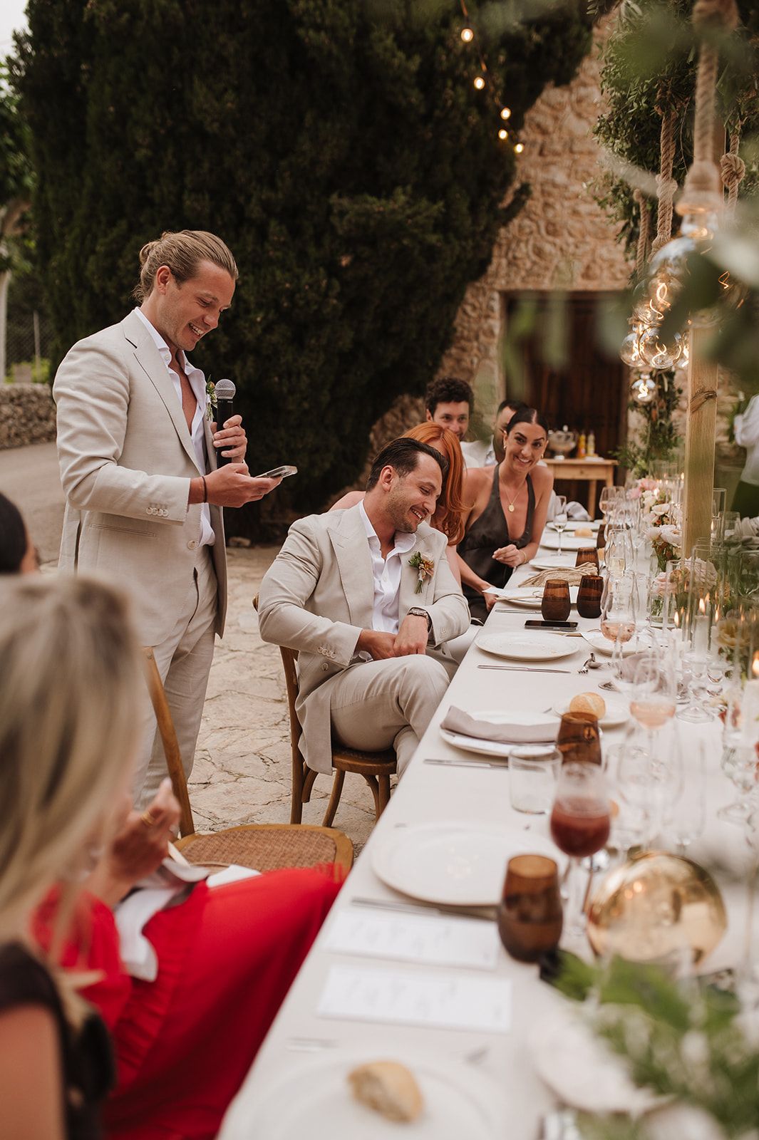 Best man giving a speech with microphone at an outdoor wedding reception dinner while guests laugh.