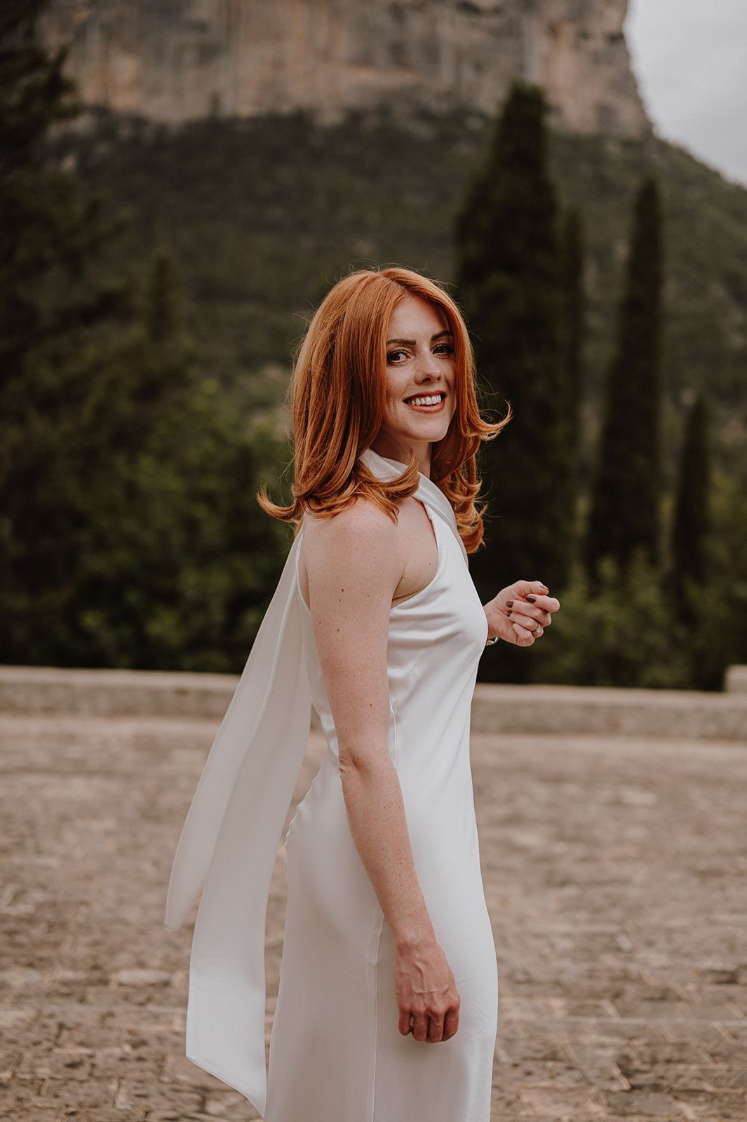 Smiling redhead bride in white halter wedding dress poses outdoors with rocky mountain scenery behind.
