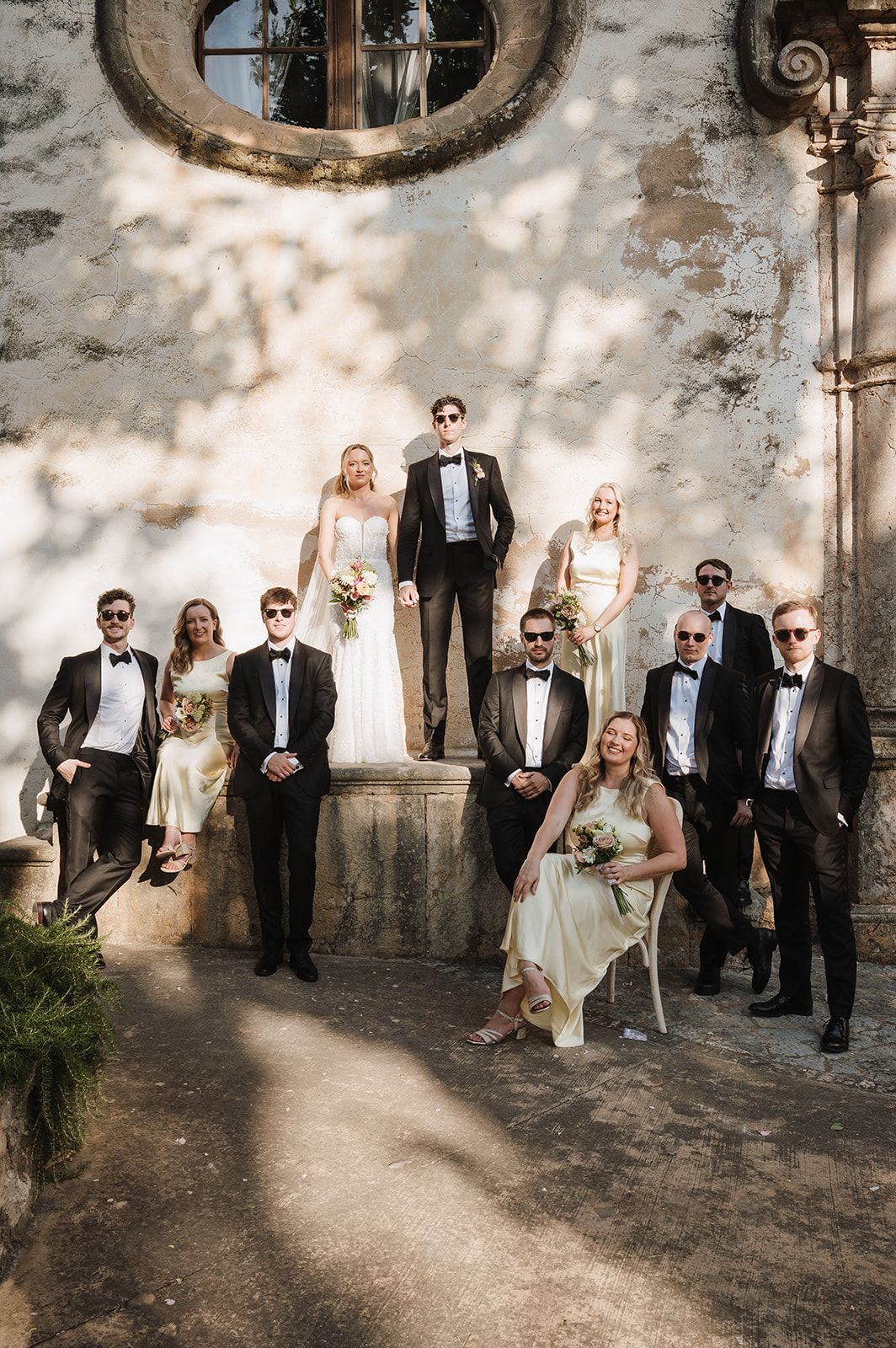 Wedding party in formal attire posing on stone steps of a rustic vintage building in sunlight.