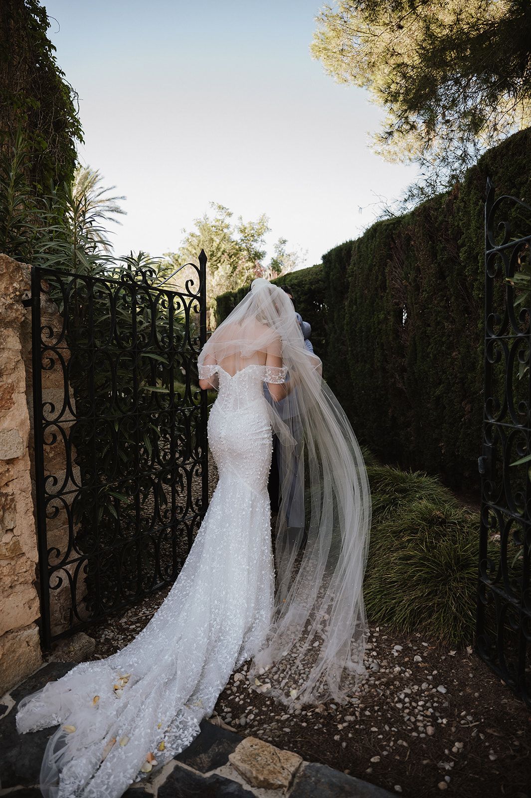 Bride in fitted lace mermaid gown with flowing veil stands at ornate iron gate in lush garden.