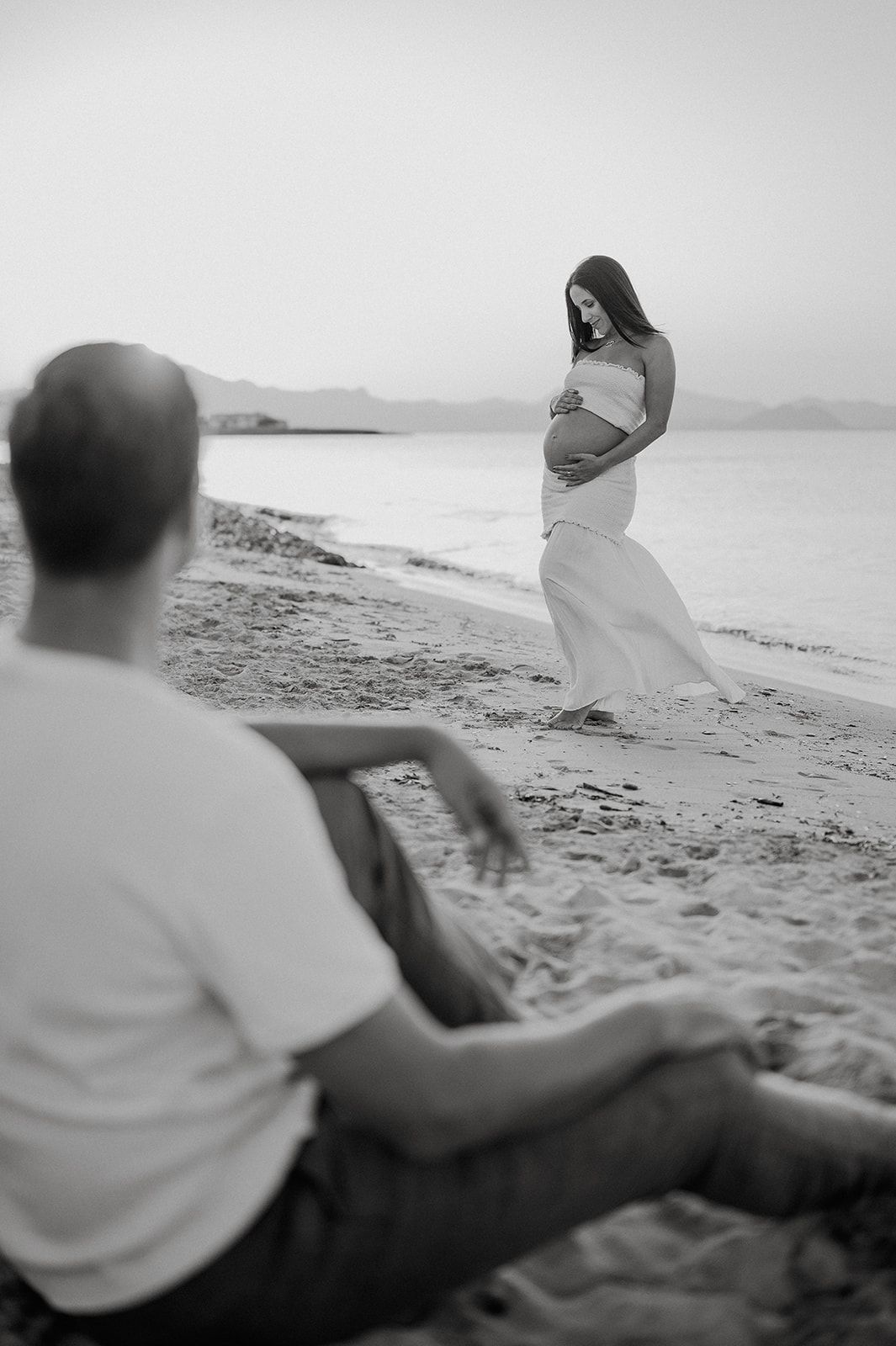 Black and white maternity photo of pregnant woman in white dress on beach with man sitting in foreground.