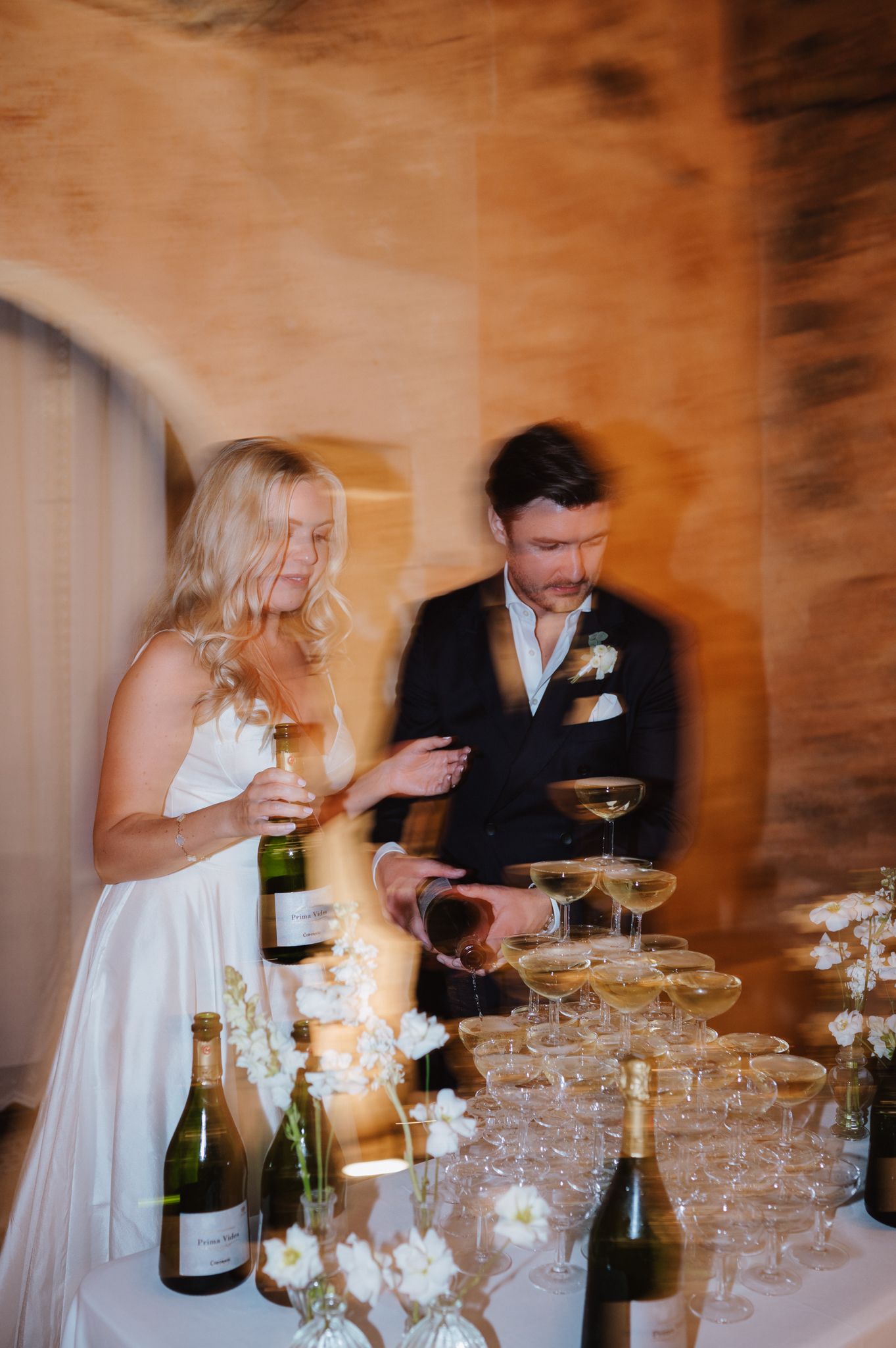 Bride and groom pour champagne tower at elegant wedding reception with floral arrangements and candlelight.