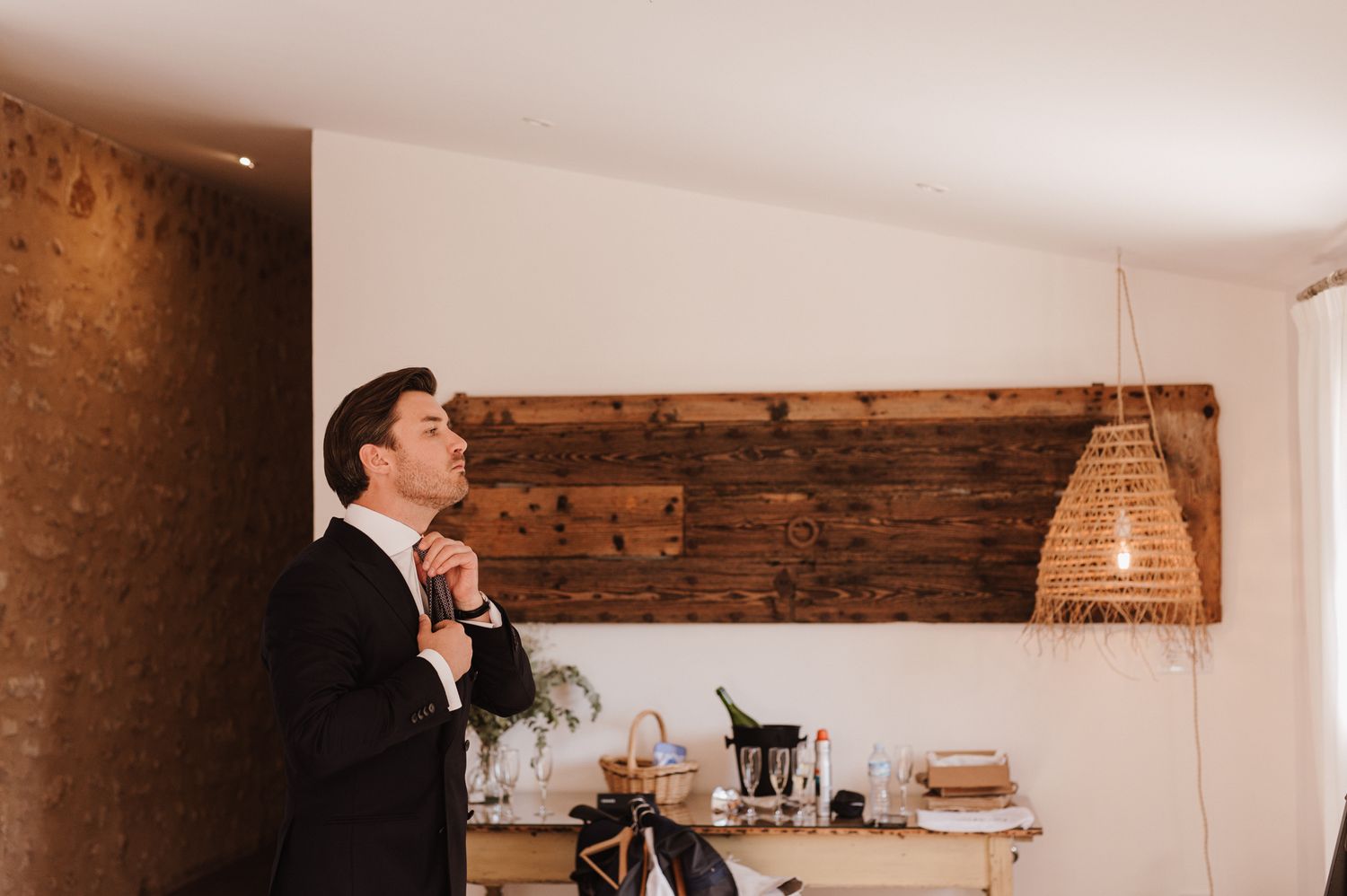 Groom in black suit adjusting his tie before wedding ceremony in rustic venue with wooden wall decor.