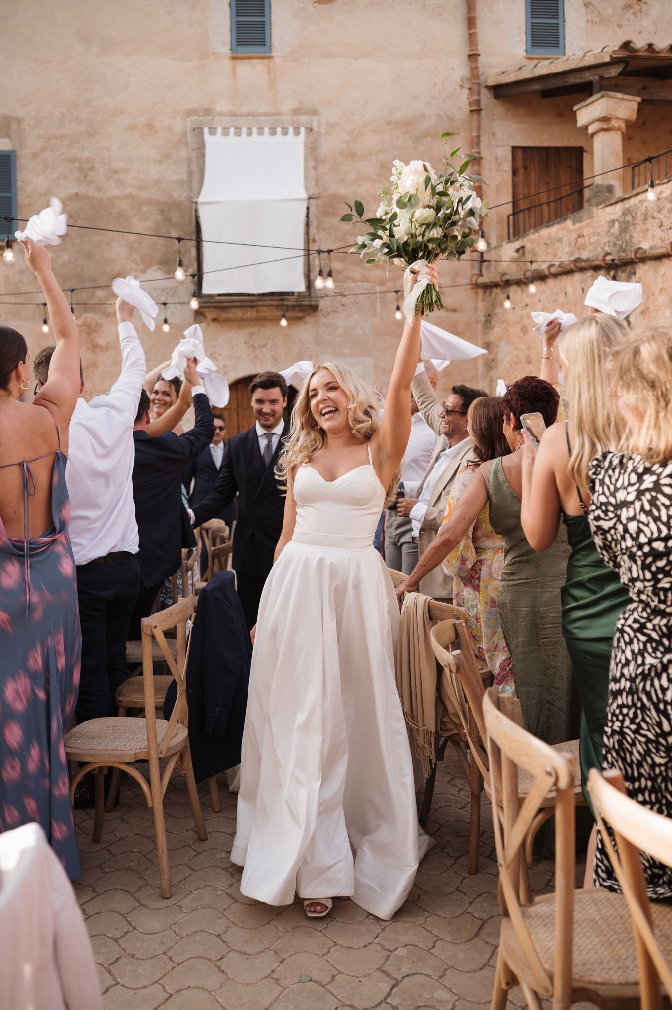 Joyful bride raises bouquet while walking through cheering guests at an outdoor rustic wedding reception.
