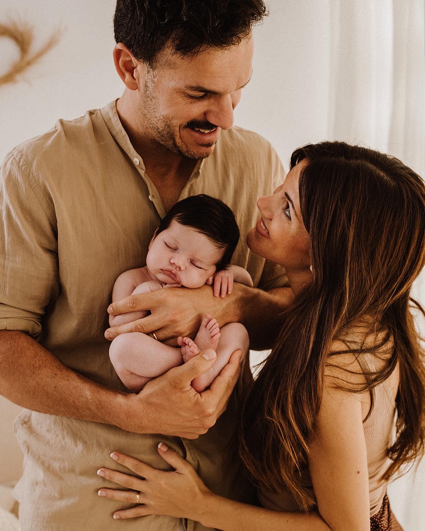 Happy couple smiling at their sleeping newborn baby with dark hair, sharing a tender family moment together.