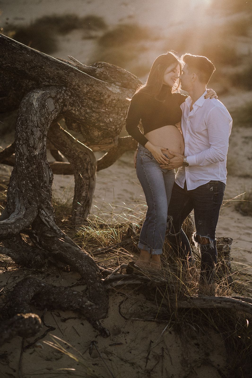Couple sharing a tender moment outdoors at golden hour, man cradling pregnant woman's belly near driftwood.