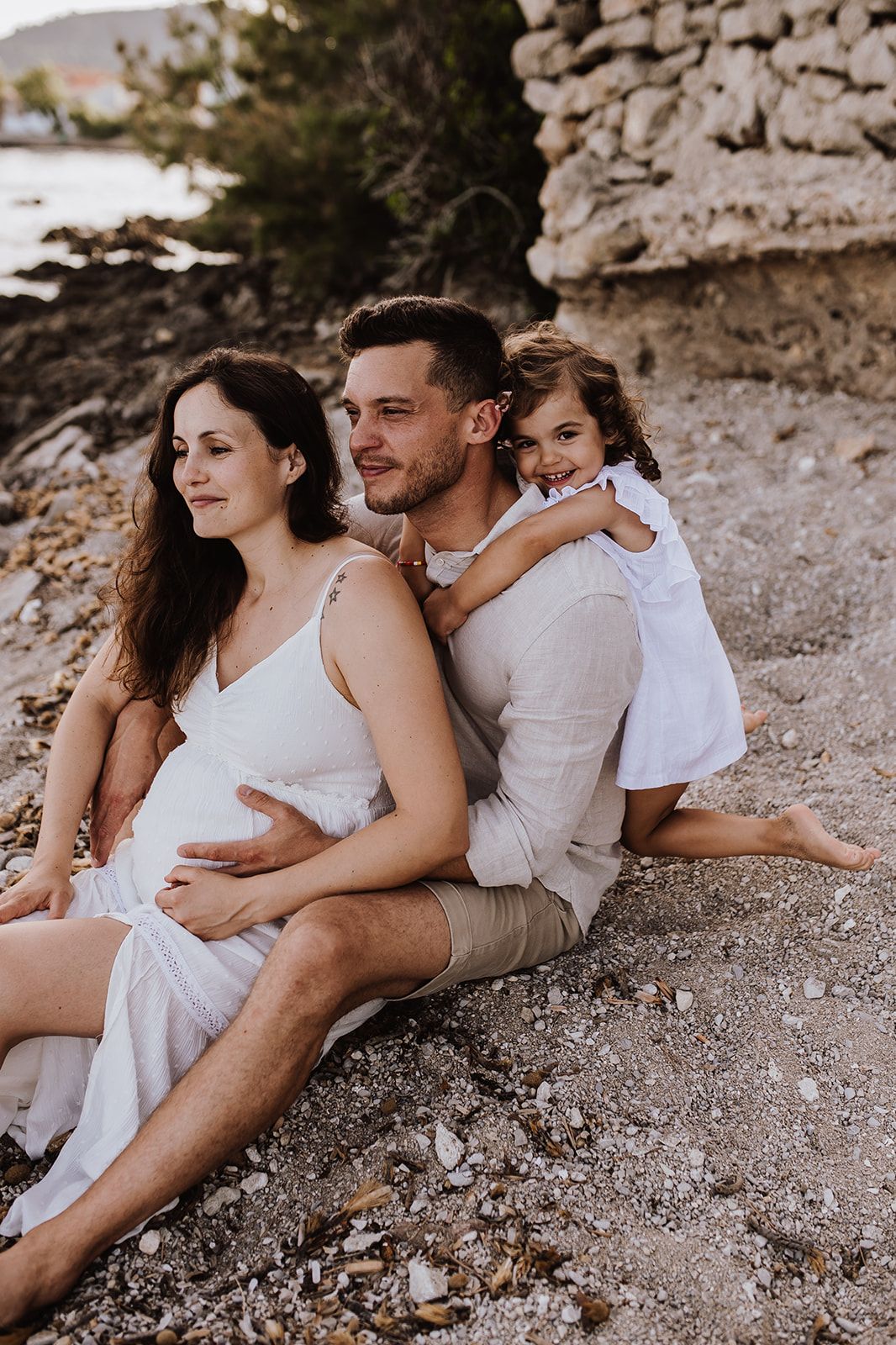 Pregnant woman in white dress sits with man and young girl on rocky beach, happy family maternity photo.