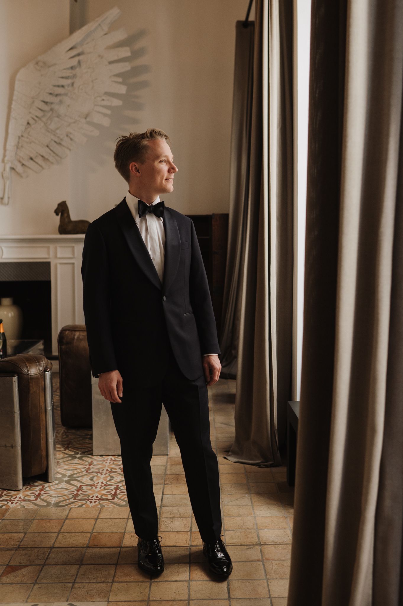 Groom in black tuxedo with bow tie standing by window in elegant room with angel wing wall decor.