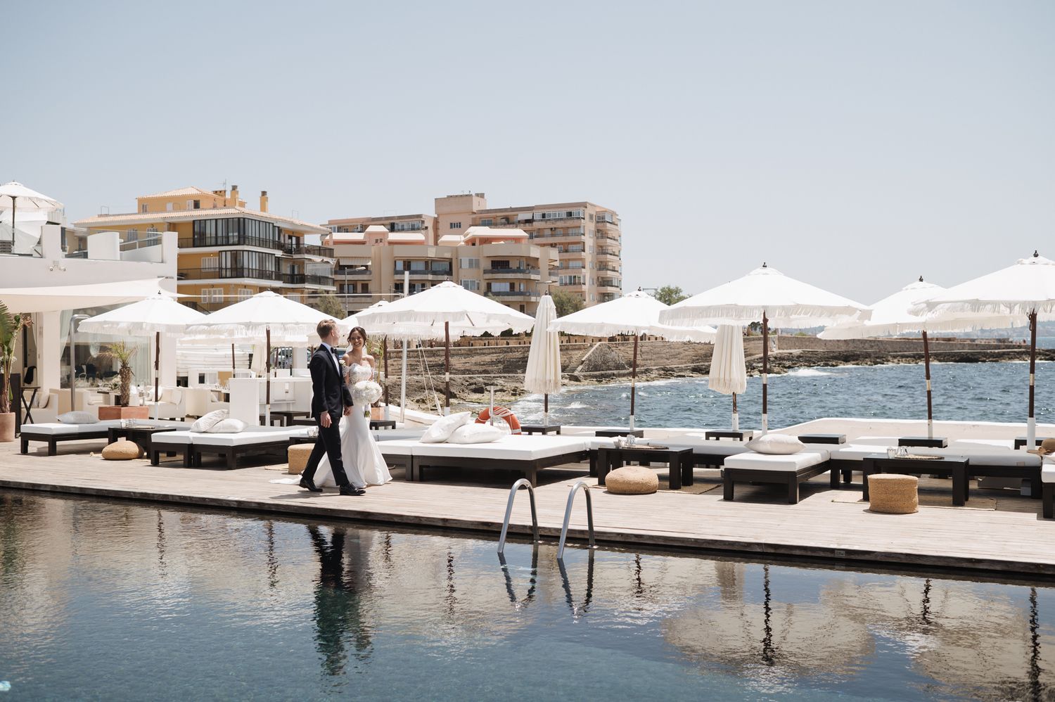Bride and groom walking along a luxury poolside venue with ocean views on their wedding day.