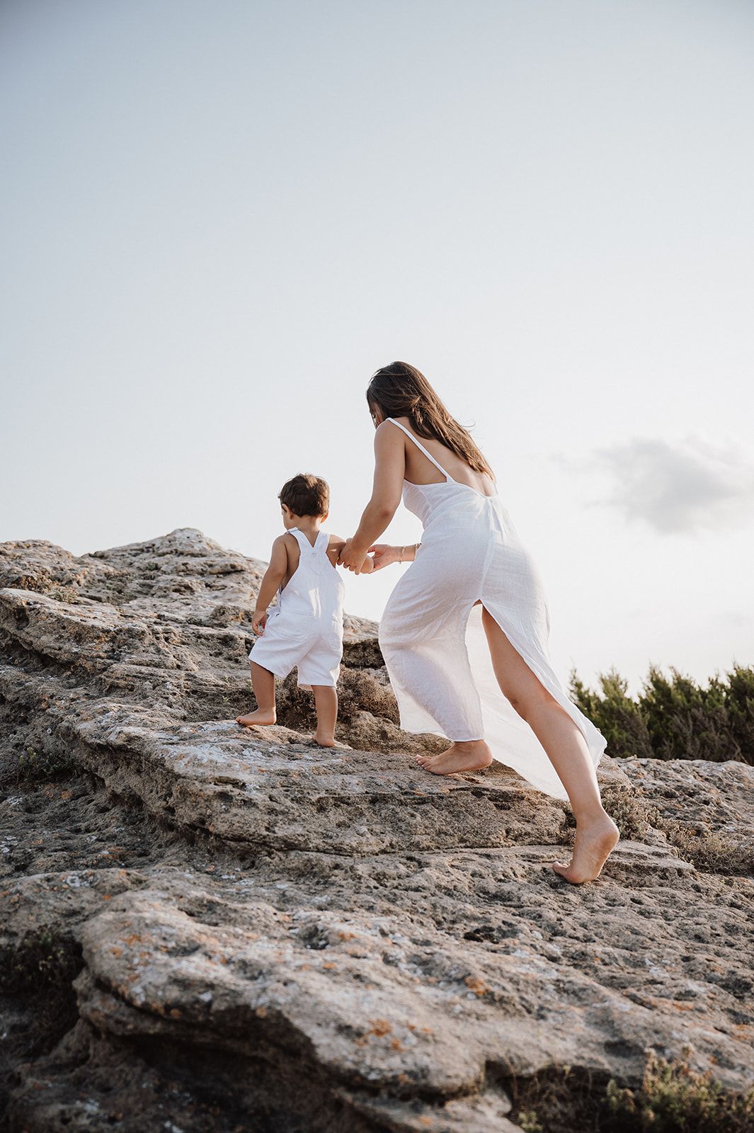 A mother in a white dress holds her toddlers hand while climbing rocky terrain at sunset.