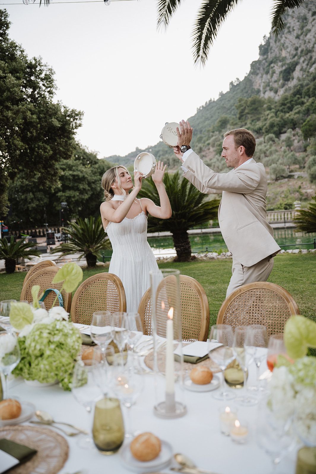 Bride and groom playfully raise tambourines over elegant outdoor wedding reception table with candles and green floral decor.