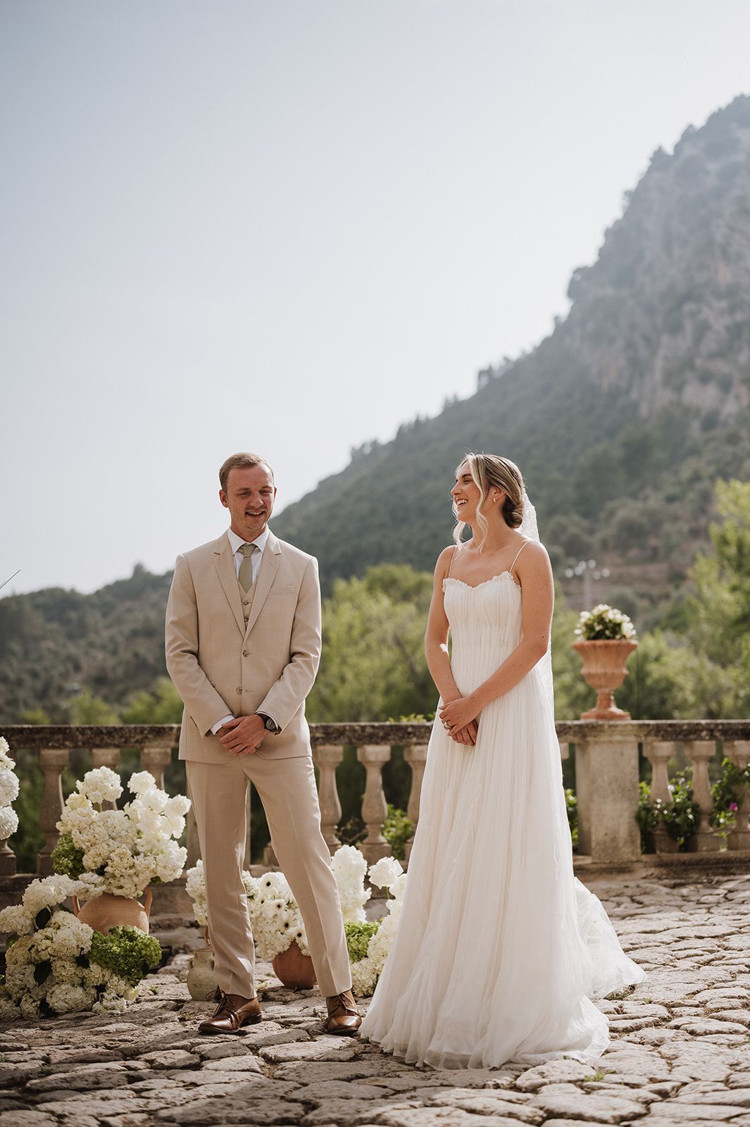 Smiling bride and groom stand together on a stone terrace with mountain views during their wedding ceremony.
