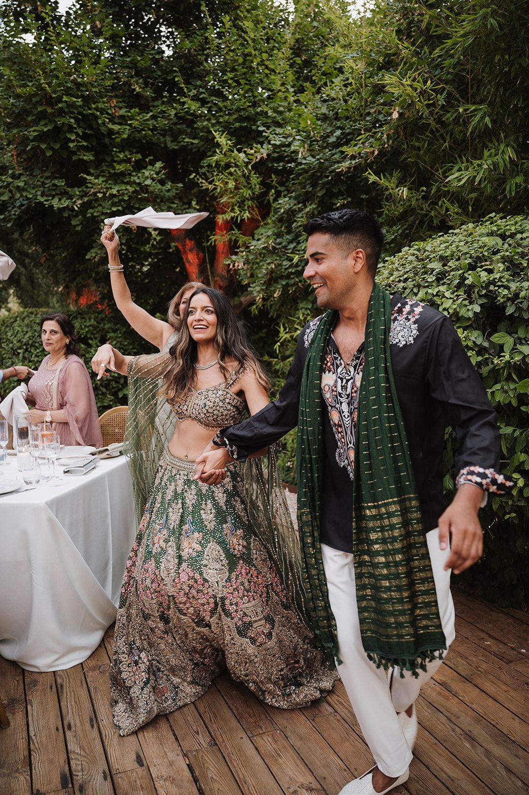 Bride in green floral lehenga dancing with groom in black sherwani at outdoor wedding reception.