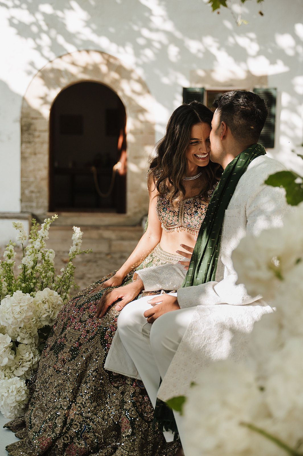 Bride and groom in Indian wedding attire sitting together laughing near white floral arrangements.