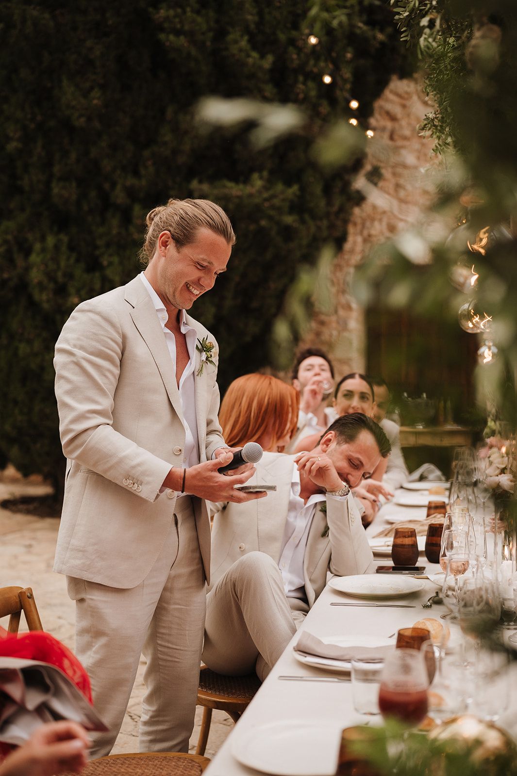 Groom in beige suit giving a speech with microphone at outdoor wedding reception.