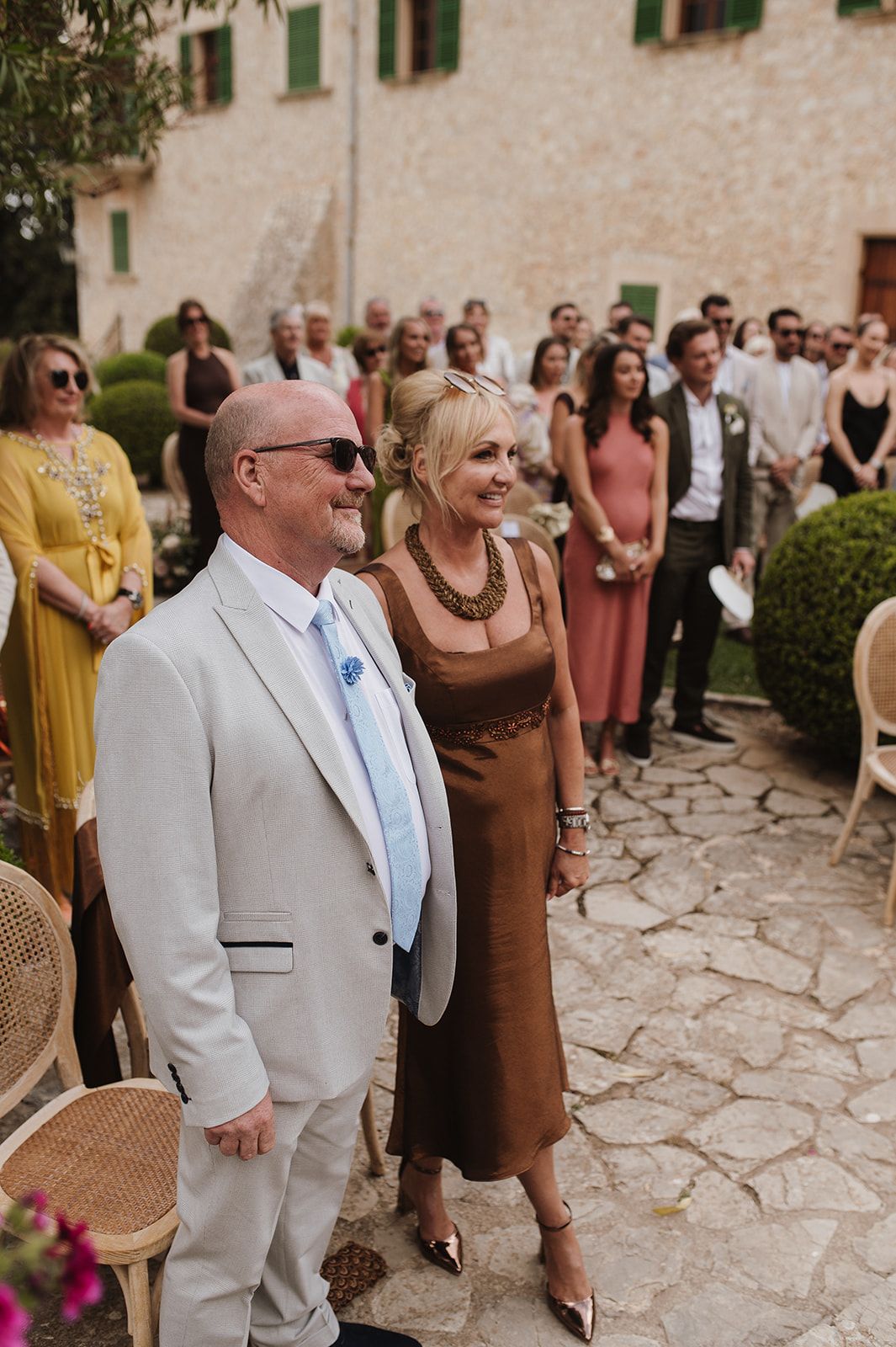 Smiling couple in beige suit and brown dress watch wedding ceremony at a rustic stone estate.