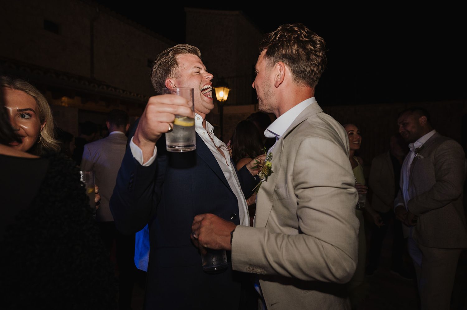 Two men in suits laughing and toasting drinks at an outdoor evening party.